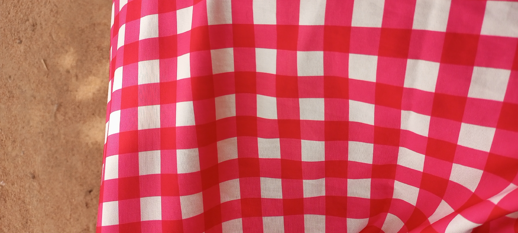 This media file is a close-up, eye-level shot of a pink and white checkered fabric. The fabric is draped, creating folds and ripples that give it texture and depth. The pattern is a classic gingham check, with alternating squares of bright pink and white. The fabric appears to be a light material, possibly cotton or a blend, and the way it falls suggests it could be part of clothing, a tablecloth, or a curtain.
The lighting in the image is even and soft, with no harsh shadows, indicating it might have been taken indoors or outdoors on an overcast day. The background, visible on the left side of the frame, is a textured, earthy-toned surface that resembles dried mud or a rough plaster wall. This provides a rustic contrast to the clean, graphic pattern of the fabric.
There is no activity or event depicted, as the focus is solely on the fabric and its pattern. No people are visible. The image is a still life, highlighting the visual appeal of the textile. There are no discernible location cues other than the possibility of the background being reminiscent of certain structures or natural formations in a place like Jalingo, Nigeria, where textures of dried earth and rough walls are common. The overall mood is simple and clean, focusing on pattern and texture. There is no visible text in the image.