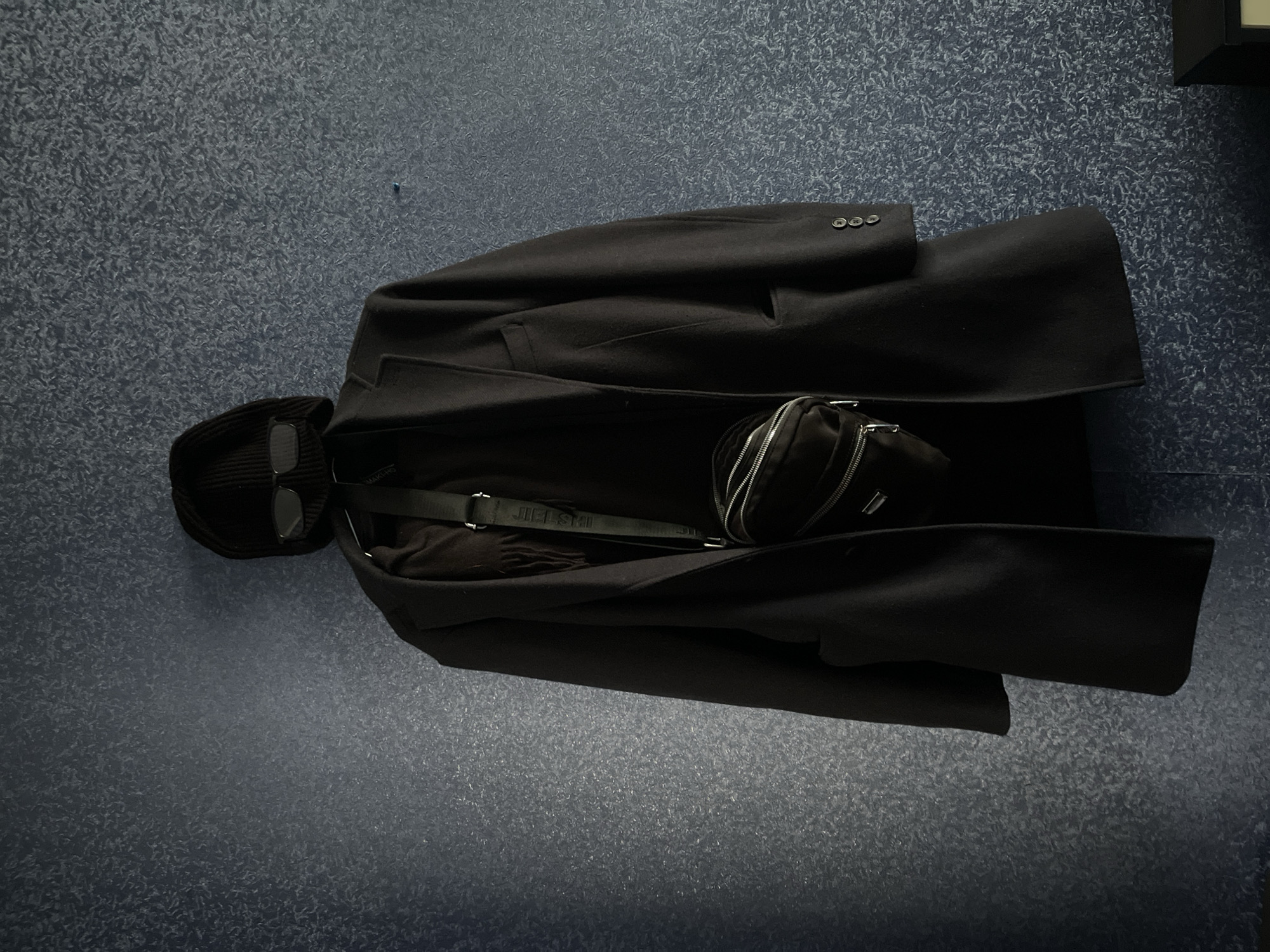 The image shows a black coat laid on a dark blue surface, alongside a black balaclava with sunglasses, and a black crossbody bag. The context doesn't suggest any specific location in Tbilisi, Georgia, and the image doesn't include people, actions, or interactions.