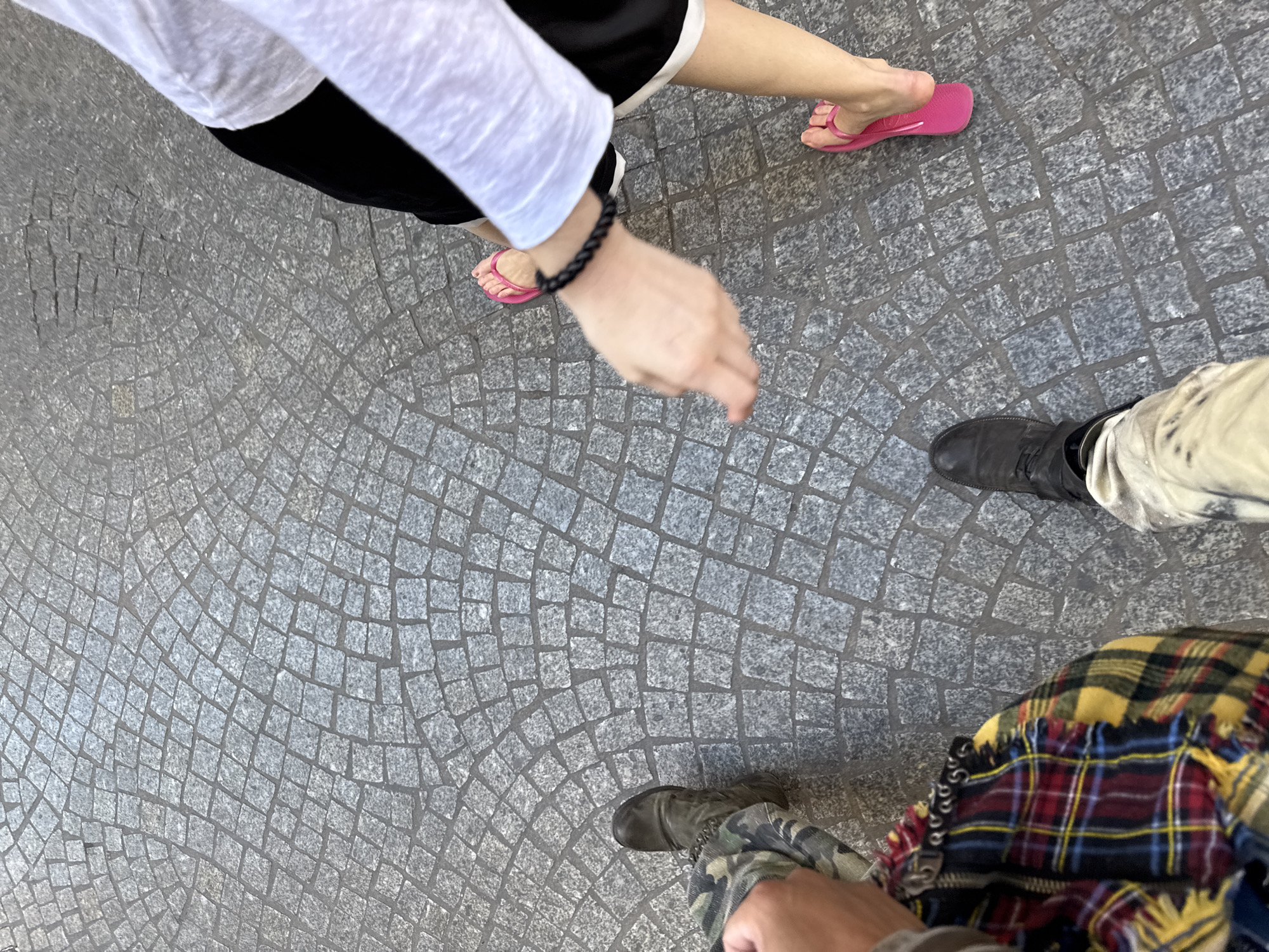 The image, taken from an overhead perspective, depicts a cobblestone pavement possibly in Paris, France. The cobblestones are arranged in a semi-circular pattern. Three individuals are partially visible. One person is wearing a white long-sleeved shirt, black shorts with white trim, and pink flip-flops. Their left arm is visible, adorned with a black braided bracelet. Another individual is wearing what appears to be off-white pants and black boots. The third person is wearing camouflage pants, olive green boots, and a tartan garment. No distinct interactions or actions are observable. The environment is composed primarily of the cobblestone pavement.