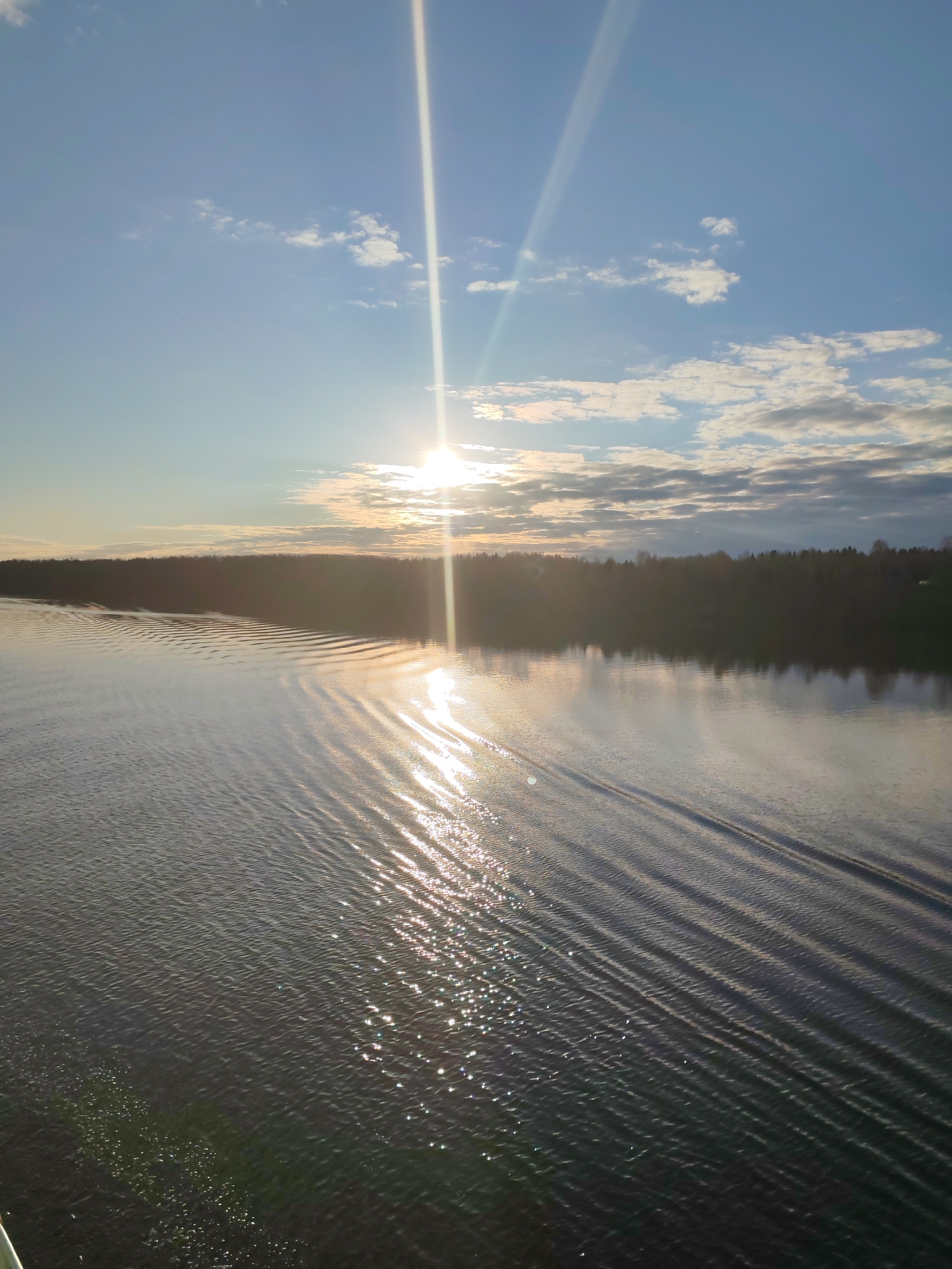 The image captures a serene natural landscape, likely a wide river or large lake, bathed in the warm light of a low-hanging sun. The scene is dominated by a vast body of water in the foreground and midground, stretching towards a distant treeline on the horizon.
The sun, intensely bright, is positioned in the upper right portion of the sky, casting a powerful glare and prominent light streaks both upwards into the sky and downwards across the water. Its reflection creates a shimmering, glittering path of light on the water's surface, extending towards the viewer. The water itself shows distinct, parallel ripples or waves, possibly a wake from a passing boat or caused by wind, which creates interesting patterns and shadows.
The sky is a clear, light blue with scattered white and grey clouds, particularly towards the right and upper areas, catching the golden light of the sun. The horizon features a dense, dark strip of land covered with trees, hinting at a forested bank.
The time of day appears to be either early morning or late afternoon, characterized by the low sun angle and the long, warm reflections. The overall mood is tranquil and picturesque. There is no visible text in the image. The location context is Раменье, Russia.