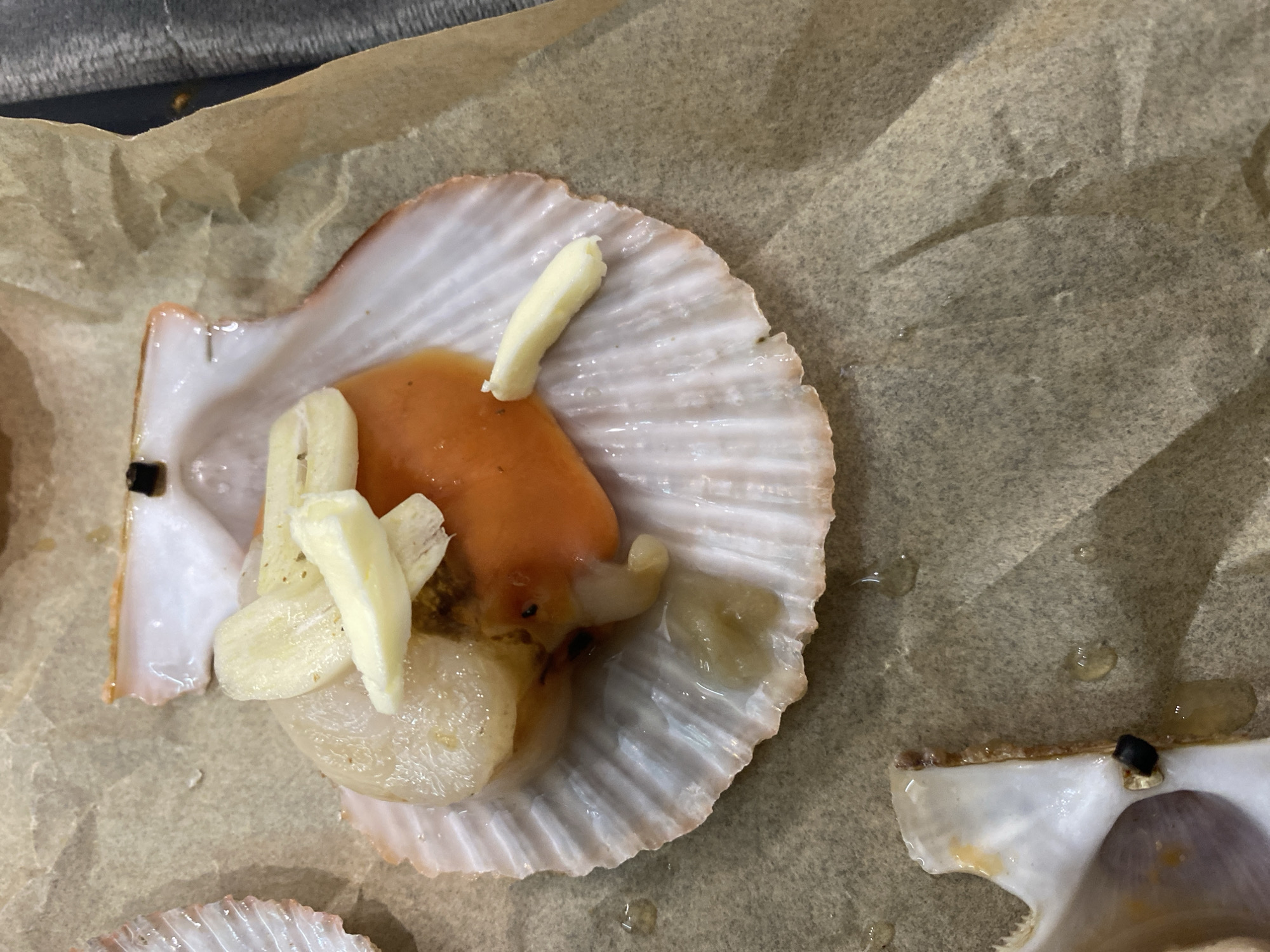 The image shows a close-up, overhead view of a raw scallop, presented in its shell and topped with slivers of garlic and butter. The scallop is situated on a piece of brown parchment paper, which is resting on a neutral-colored, textured surface, possibly a countertop or baking sheet. The scallop itself is plump and appears fresh, with its characteristic orange coral and white adductor muscle visible. The shell is iridescent and ribbed, with hints of pink and orange along its rim. Small droplets of liquid, likely from the scallop or butter, are scattered on the parchment paper. The lighting is bright, emphasizing the textures and colors of the food. There is no indication of any specific location within Novorossiysk, Russia, or any people or actions taking place. The scene is static and focuses solely on the prepared scallop.