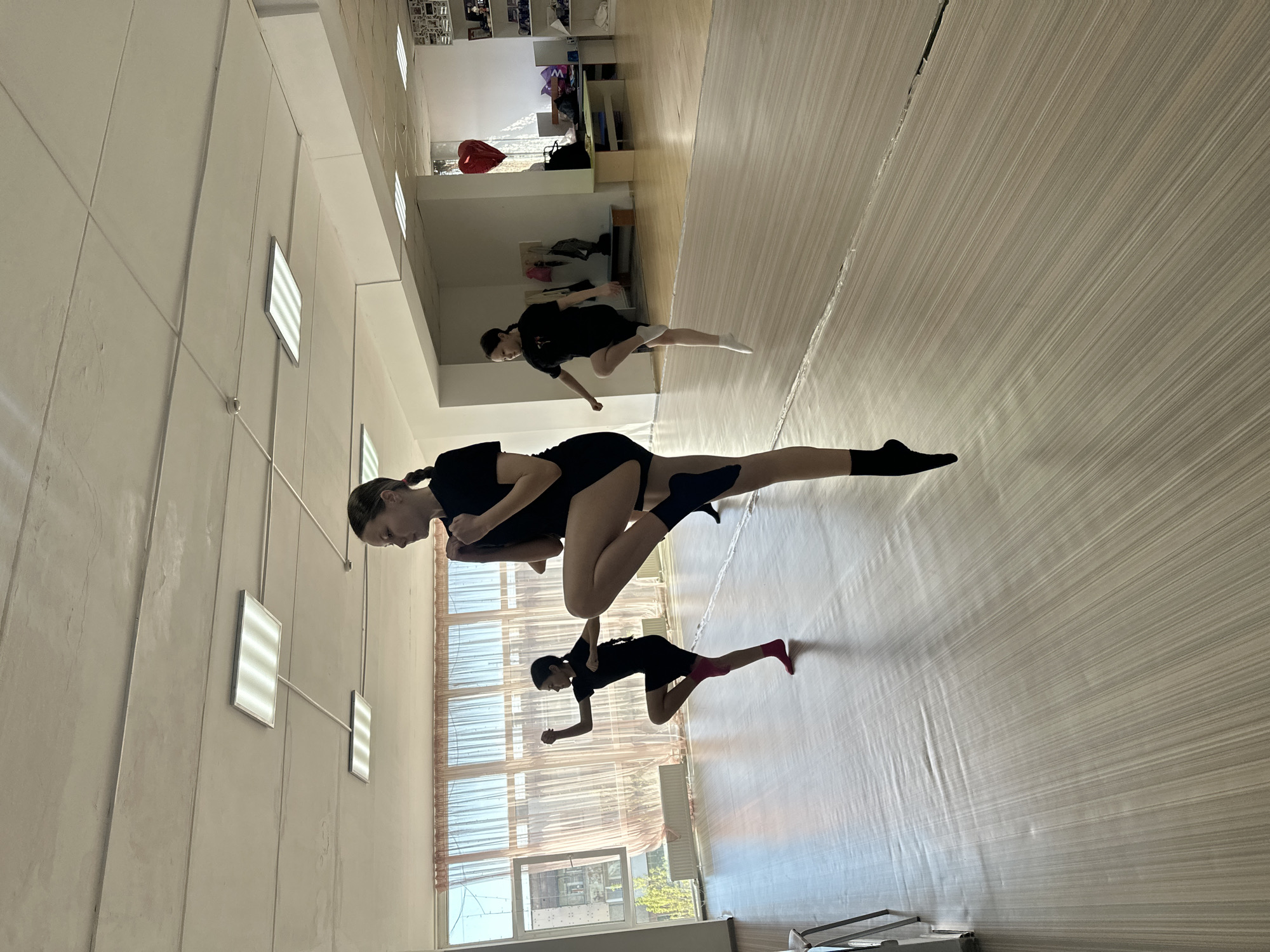 The image shows three dancers in a studio, likely in Votkinsk, Russia. The studio has a light-colored wood floor and white walls. There are rectangular fluorescent lights on the wall. A large mirror is visible on one side of the room, reflecting the scene. One dancer is positioned in the foreground in a contorted pose, seemingly supported by another. A third dancer is in the background. All dancers are wearing black leotards. One is also wearing pink socks and one has black socks on. The foreground dancer has tape around her knee. The dancers appear to be practicing or performing. Outside the window, trees are visible, suggesting a daytime setting.