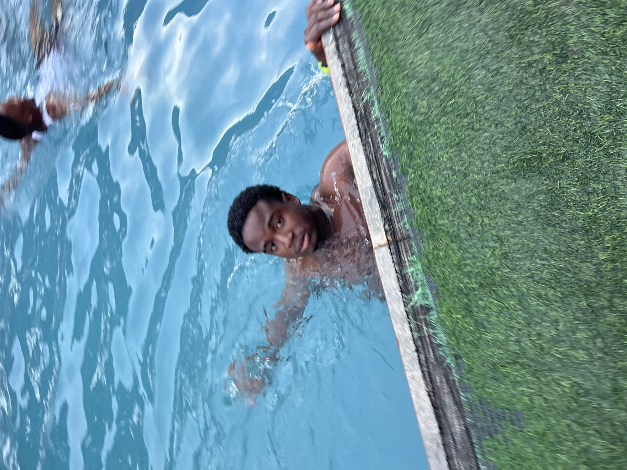 This image captures an outdoor recreational scene, likely in Nkpor, Nigeria, featuring two individuals in a body of water, probably a swimming pool. The primary subject is a dark-skinned young man, positioned in the right half of the frame, partially submerged in bright blue water. He is looking directly at the camera with a relaxed expression, his head and torso visible above the water, while he holds onto the concrete edge of the pool with his right hand. Water droplets cling to his skin and short, dark hair.
In the upper left quadrant, another person is partially visible, mostly submerged in the water and wearing a white top. The water exhibits clear ripples and reflections, suggesting it is daytime with bright conditions. Adjacent to the pool's edge on the right side of the image, there is an area covered with vibrant green artificial turf or grass, bordered by a dark mesh fence or netting. The scene conveys a casual and enjoyable atmosphere, indicating a leisure activity such as swimming. No text is visible in the image.