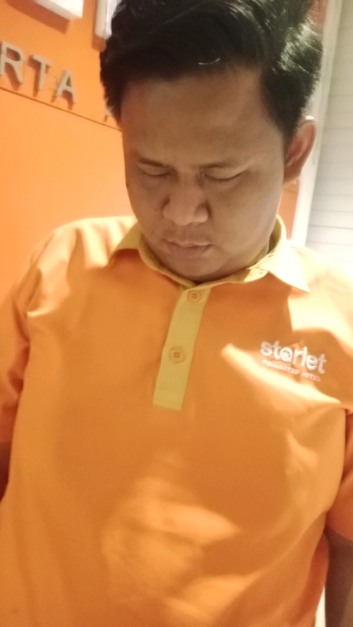 The image shows a close-up of a person wearing an orange polo shirt with a yellow collar and button placket. A white logo that reads "starlet" and "CONNECTED HOTEL" is visible on the right side of the shirt. In the background, a portion of an orange wall is visible with white lettering that appears to spell out "RTA." This lettering and the orange wall suggest the location is likely within the city of Tangerang, Indonesia, potentially at a business or establishment with this signage. The person is looking downwards, and their face is partially obscured. The lighting suggests an indoor setting.