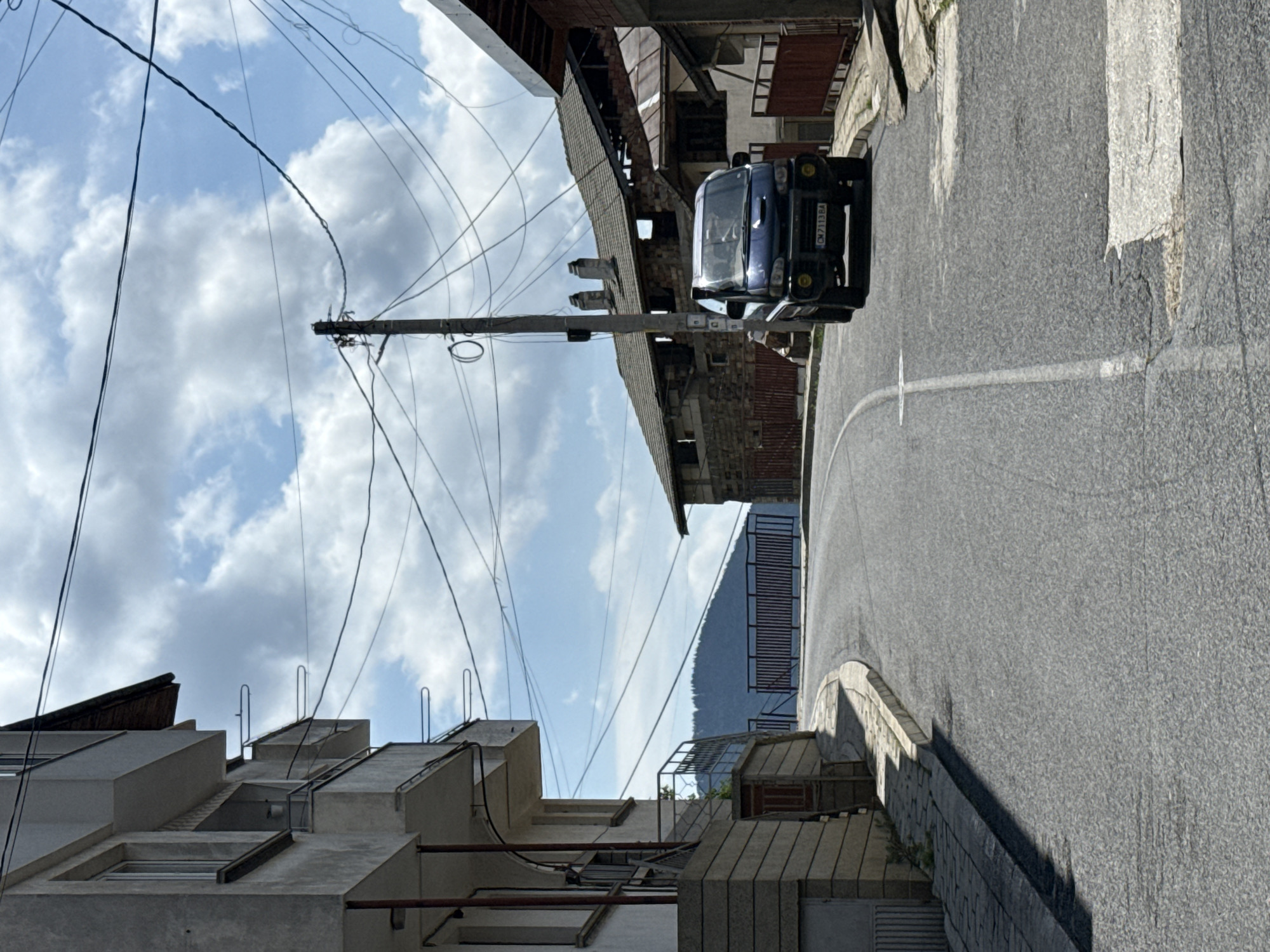 The image, originally rotated, shows a street scene under a partly cloudy sky. An asphalt road, marked with a white center line, curves to the left in the distance. A dark blue minivan or small truck is parked on the right side of the road, facing the viewer, with its license plate "CM 7113 BA" clearly visible.
A prominent wooden utility pole, heavily laden with numerous black wires, stands in the left-center foreground. On the left side of the road, there is a multi-story building with a modern, blocky, concrete architectural design, featuring several windows and roof access ladders or railings. On the right side and in the background, older structures with exposed brick, corrugated metal, and wooden elements are visible. In the distant background, a large, dark, tree-covered mountain or hill rises against the sky. The scene is brightly lit by sunlight, casting shadows, indicating a clear day with scattered white cumulus clouds. No people are visible.