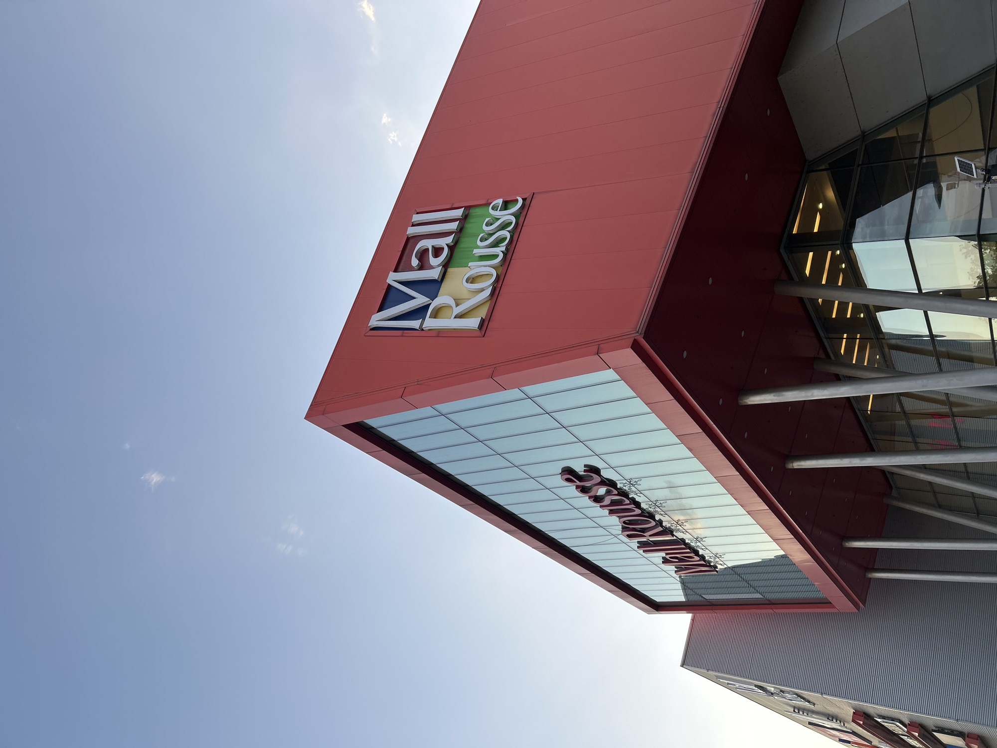 The image shows the facade of the Mall Rousse in Ruse, Bulgaria, under a clear blue sky with a few wispy clouds. The mall has a modern architectural design, with a dominant bright red upper section that angles sharply towards the sky. This red section features a prominent logo with the words "Mall Rousse" in a multi-colored block font, with "Mall" in blue and white, and "Rousse" in yellow and green. Below this, a large expanse of reflective glass windows covers the lower part of the building, reflecting the sky and the surrounding environment. Several cylindrical metal pillars support the overhang of the red section, leading down to the entrance area which is also largely glass. To the left of the red section, a gray, corrugated metal building is partially visible, also appearing to be part of the mall complex. The perspective is from a low angle, looking up at the building. There are no people visible in the image, and the scene appears to be quiet and unoccupied.