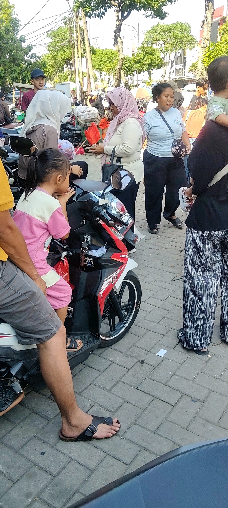 The image depicts a bustling outdoor scene in Cibatu, Indonesia, likely a market area given the presence of various people and a parked motorcycle. A young girl in a pink and white shirt is seated on a red and black scooter, looking to her left. Beside her, an adult male wearing grey shorts and black sandals is partially visible, sitting on the scooter as well. In the background, several individuals are engaged in various activities. A woman in a light blue shirt and black pants stands with her arms crossed, while another woman in a pink headscarf and a light-colored top converses with someone out of frame, holding a red flower or object. A person wearing a black hijab and patterned pants walks away from the camera. The ground is paved with interlocking grey bricks. Trees line the background, with a building and overhead wires suggesting an urban environment. The lighting indicates it is daytime.
