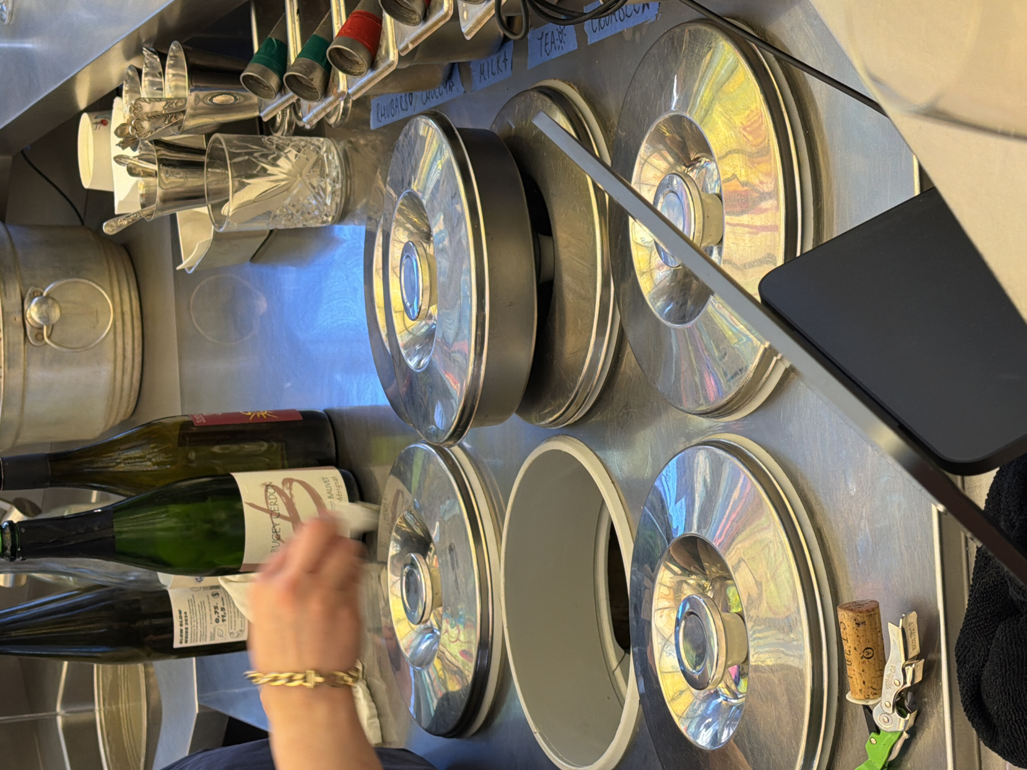 Here is a detailed description of the image:
**Environment:** The scene appears to be a workstation, possibly in a restaurant or bar. The surfaces are stainless steel, and the overall impression is clean and utilitarian. The lighting is bright and reflective.
**Objects:**
* Several metal containers/drums or reservoirs, possibly for ingredients or beverages.
* Bottles of wine/liquor are visible, including one labeled "BUGEY CERDON".
* Utensils and glasses are stored in holders.
* A digital tablet or screen is present, possibly for order management or payment.
* A corkscrew and cork.
* Small labels with words like "RHUBARSD, CMLLYS MILKA TEAR COUMOLUN" are present.
**People:**
* Part of a person's arm and hand is visible. The person is wearing a gold bracelet and cleaning one of the wine bottles with a cloth.
**Actions:**
* Cleaning of a wine bottle.
**Interactions:**
* A person is interacting with objects and cleaning the bottle.
**Possible Location:** The labels contain the letters 'MILKA TEAR COUMOLUN', which could be an acronym. It's not immediately apparent if the location is London, United Kingdom, unless there are specific identifiers not discernible in the image.