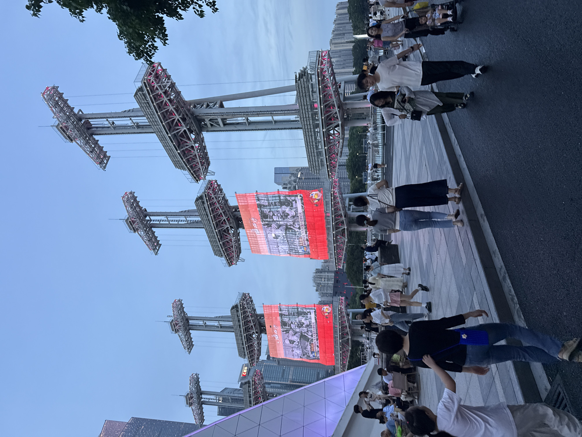 The image captures a bustling scene at dusk in Tianhe District, China, likely at an outdoor amusement park or entertainment venue. Dominating the frame are several towering structures that appear to be part of a thrill ride, with their intricate metal frameworks illuminated by red lights. Suspended between these structures are large, rectangular red screens displaying images. The screens show what seem to be historical or celebratory scenes, with some text visible, including what appears to be Chinese characters.
A diverse group of people are seen walking along a paved walkway in the foreground and middle ground. They are dressed in casual summer attire, suggesting a warm evening. Some are walking in groups, while others are alone. The atmosphere appears lively and social, with people seemingly enjoying their time. The lighting suggests it is either early evening or late afternoon, with the sky a pale blue transitioning to a darker hue. The setting includes modern high-rise buildings in the background, typical of a large urban environment in China. The overall impression is one of a vibrant public space with a popular attraction drawing a crowd.