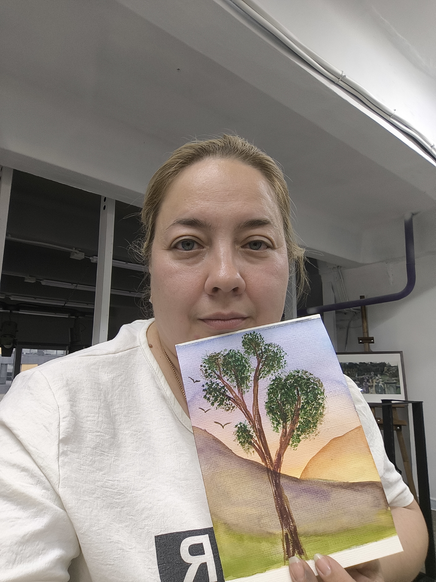 The image depicts a woman holding a watercolor painting in front of her. The painting features a tree in the foreground against a backdrop of hills and a sunset. The woman has fair skin, blonde hair, and is wearing a white t-shirt. She appears to be indoors, with a white ceiling and structural elements visible in the background. An easel with a framed picture is visible behind her. The location is potentially a studio or art space. Without further evidence, it is not possible to definitively confirm that this scene is in Moscow, Russia.