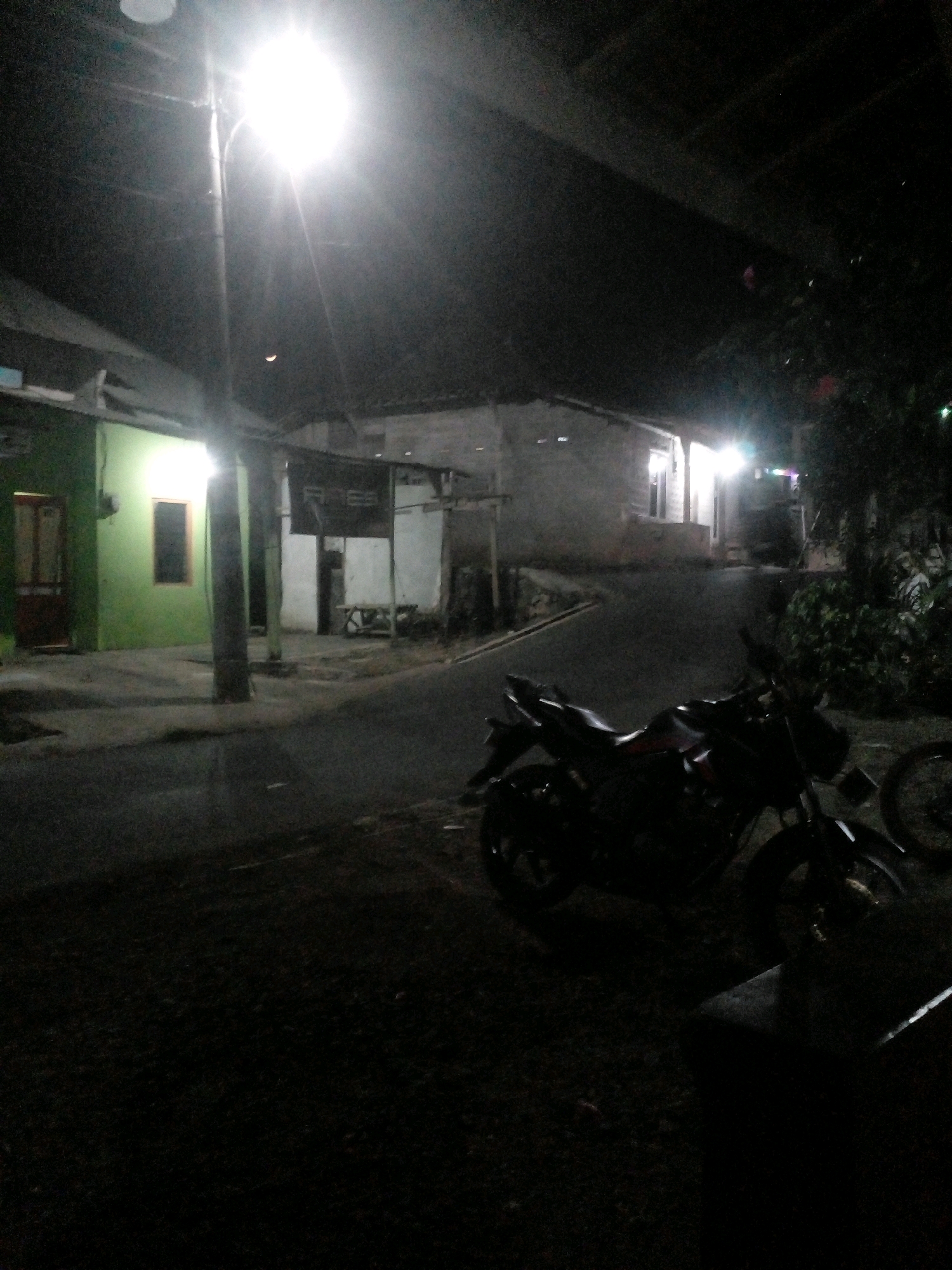 The image depicts a dimly lit street scene at night, likely in Pungangan, Indonesia. A bright streetlamp illuminates the upper left portion of the frame, casting long shadows. In the foreground, a black motorcycle is parked on the asphalt road. To the left, a green building with a brown door and a small rectangular window is visible. Adjacent to it, a white building with a sign that reads "RAGS" and a wooden bench is also present. The background shows more structures and dense foliage, characteristic of a residential area in a tropical climate. The crescent moon is faintly visible in the dark sky.