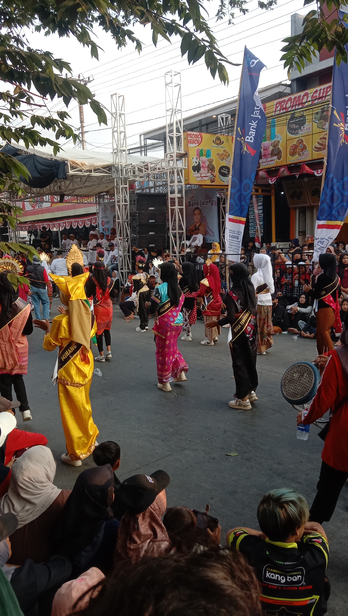 In Jatisawit, Indonesia, a street performance or parade is underway, with several young people dressed in vibrant, traditional costumes. The individuals in the foreground are wearing brightly colored garments, including a gold outfit with a distinctive headpiece and a pink dress with intricate patterns. Some participants are adorned with sashes bearing place names such as "MALUKU" and "NUSA TENGGARA."
The scene is set on a paved street, with a crowd of spectators gathered on either side, many seated on the ground. In the background, a stage structure with large speakers and scaffolding is visible, suggesting a musical or theatrical performance. Billboards with advertisements, including one for "Bank Jateng," are also present. The event appears to be taking place outdoors, with tree branches framing the top of the image and the sky visible above. The overall atmosphere suggests a cultural celebration or festival.