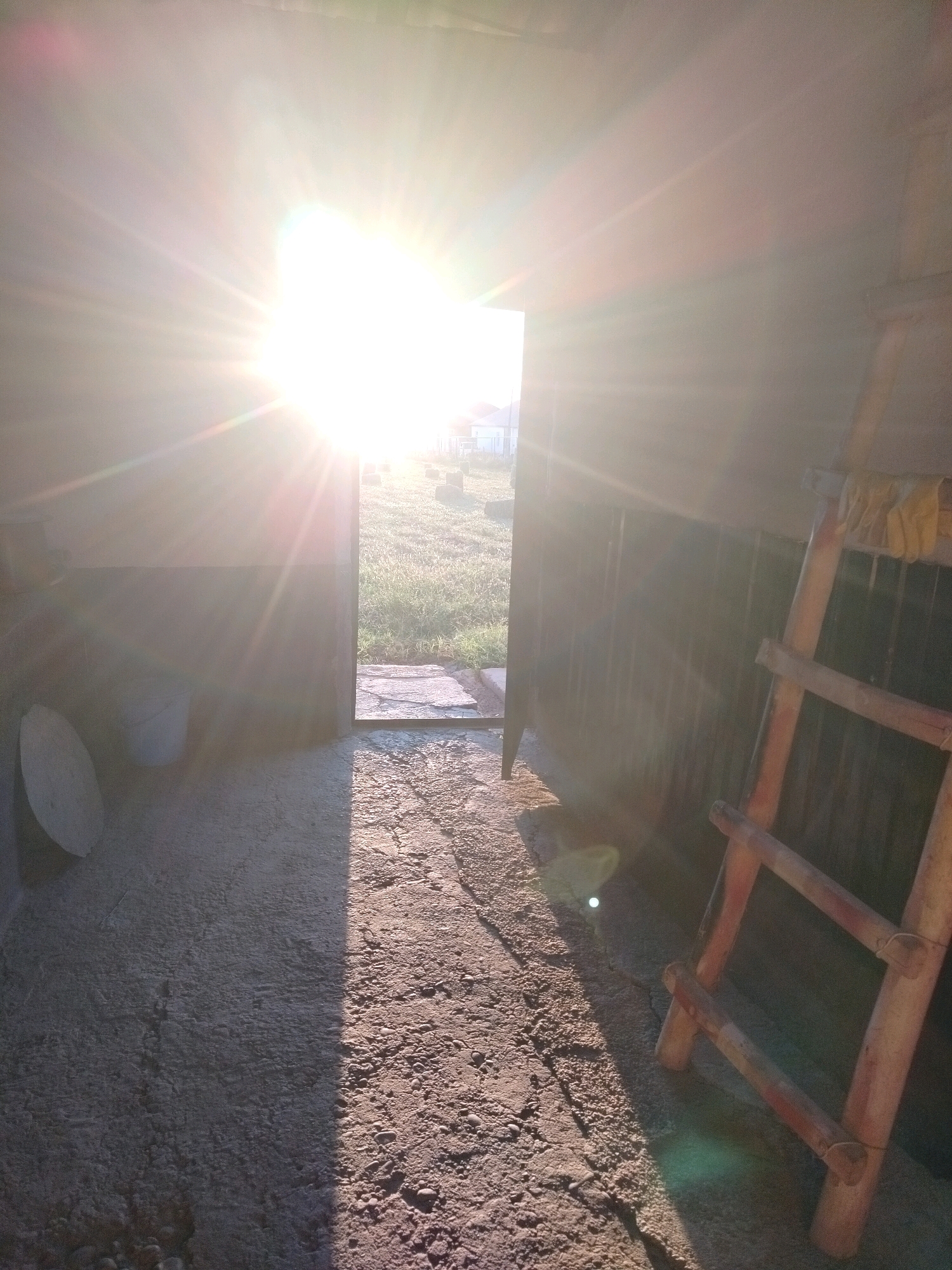 The image depicts the interior of a dimly lit structure, possibly a shed or garage, with a bright, sunlit doorway. The sun is positioned directly in front of the doorway, creating a lens flare and casting strong rays of light into the interior. The floor inside is uneven and appears to be made of concrete and gravel. To the left, a white bucket and a round, flat object are visible against a wall. To the right, a wooden ladder leans against a dark, corrugated metal wall. Beyond the doorway, a grassy area with scattered round objects, possibly bales of hay, is visible, along with a faint suggestion of buildings in the distance. The overall impression is that of a rustic, rural setting bathed in the intense light of sunrise or sunset. There are no people present in the scene. The location is identified as a property in the city of Kölkent, Kazakhstan.