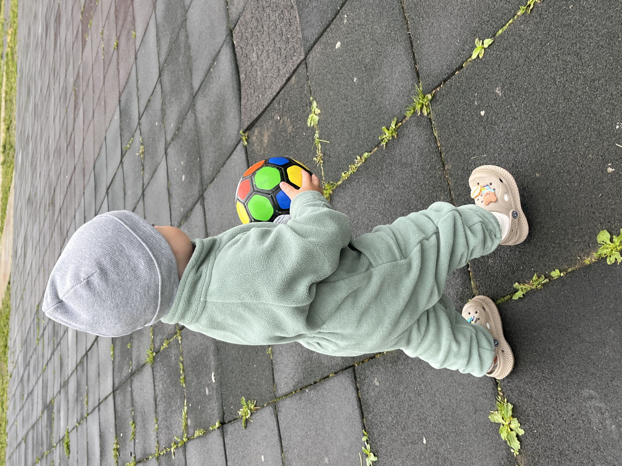 An overhead shot captures a young child holding a colorful soccer ball on an outdoor surface in Chaikovsky, Russia. The child is dressed in a light grey ribbed beanie and a mint green fleece jumpsuit or tracksuit, suggesting cool weather. On their feet are light beige/tan Crocs with white socks peeking out, decorated with small, colorful charms.
The child is positioned centrally in the frame, viewed from the head down to the feet, holding a black, red, blue, green, and yellow paneled soccer ball with both hands. Partially visible on one of the black panels of the ball is the word "DESIGNED".
The setting is an outdoor area paved with dark grey rubber tiles, possibly a playground or sports court. Weeds and small patches of green grass are growing through the seams and cracks of the tiles, indicating natural growth and an outdoor environment. A patch of vibrant green grass is visible in the background to the left. The lighting is soft and diffused, characteristic of an overcast or cloudy daytime. The image provides a candid, top-down view of the child engrossed with the ball. A logo is visible on the sole of the right croc.