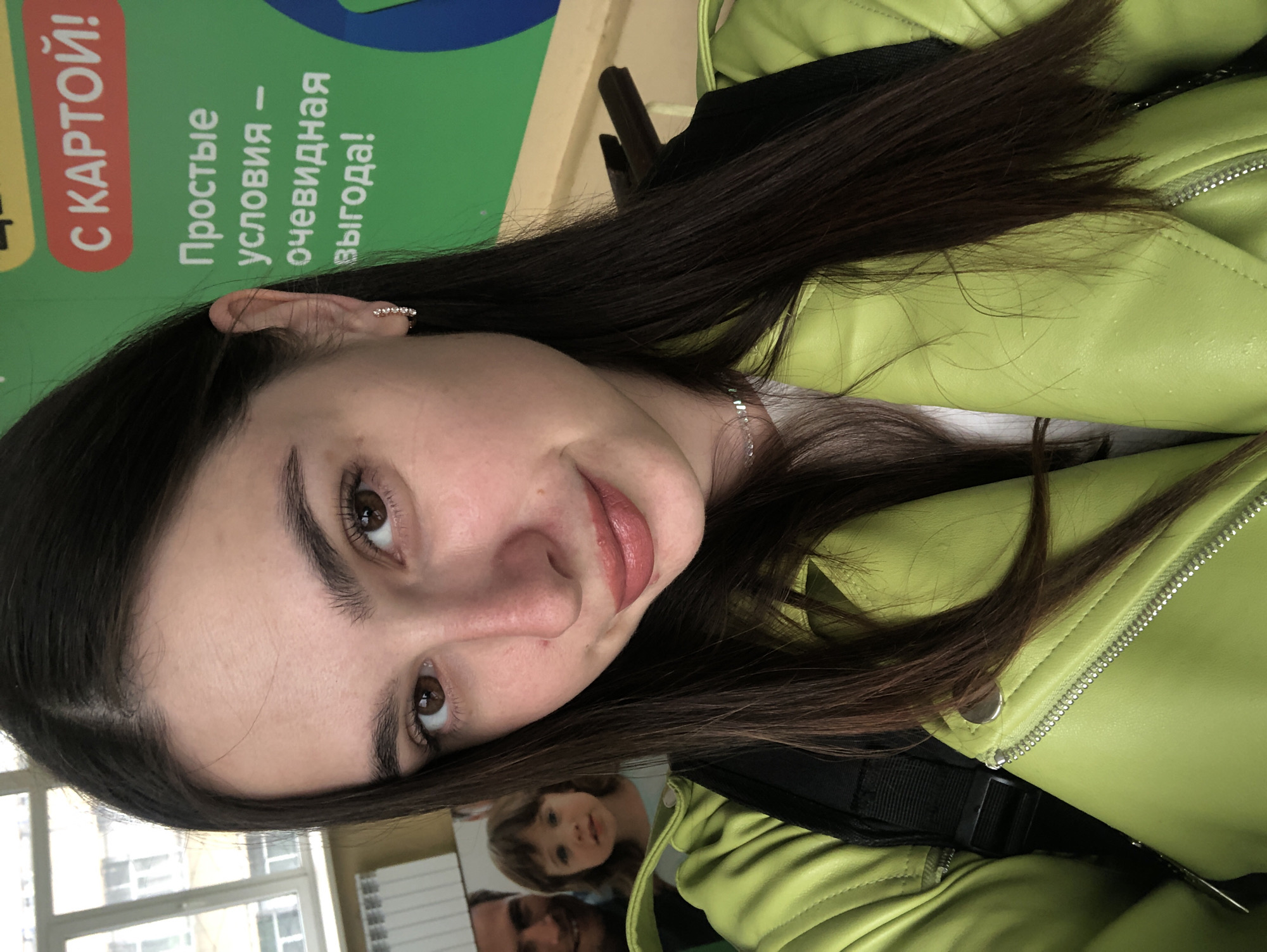 The media file is a close-up, tilted selfie of a young woman. She has long, dark brown hair and brown eyes. She is wearing a bright lime green jacket with a zipper and a silver button. She is also wearing subtle earrings and a delicate silver necklace.
In the background, a green sign with white text is visible, which appears to be an advertisement. The text on the sign is in Russian and reads: "С КАРТОЙ! Простые условия — очевидная выгода!" (With a card! Simple conditions - obvious benefit!). To the left, through a window, some buildings and possibly a street can be seen, suggesting an indoor setting with a view to the outside. There is also a reflection visible on a shiny surface, showing a glimpse of another person and a child, possibly a family photo.
The lighting suggests it might be daytime, but the overall mood is informal and candid, likely taken by the subject herself. The orientation of the image suggests it was taken in portrait mode. The subject's facial expression is neutral to slightly smiling. The location context is Chaikovsky, Russia.