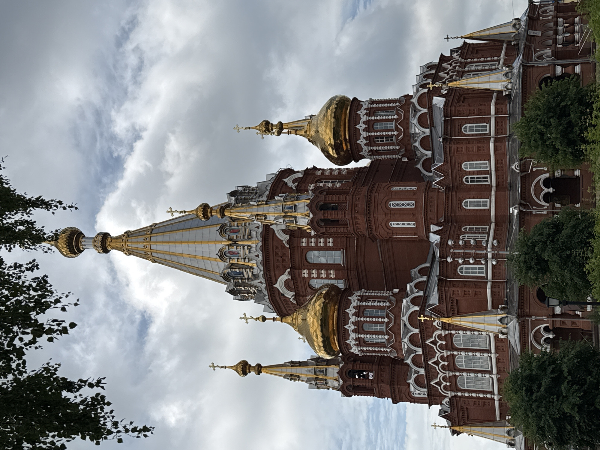 The image shows a large, red brick Orthodox church with golden domes and crosses under a cloudy sky. The church has multiple towers and intricate white detailing around the windows and rooflines. Green trees and foliage are visible in the foreground, obscuring the lower portion of the building. A streetlamp is also present among the trees. This is the Holy Trinity Cathedral, located in Izhevsk, Russia. There are no people visible in the image.