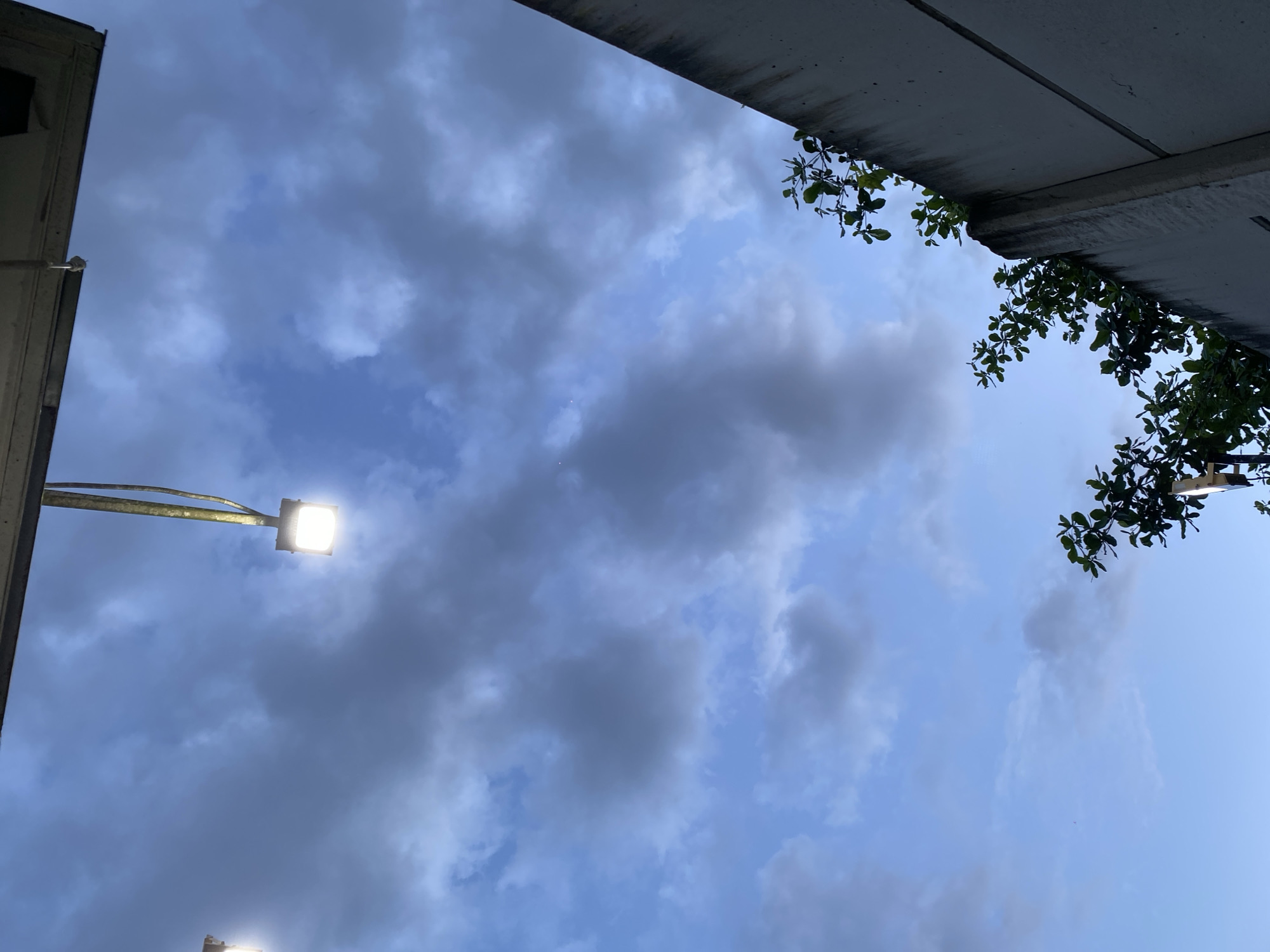 The image captures a cloudy sky with two illuminated outdoor lamps, one on the left and another on the right. A concrete wall runs along the right side of the frame, topped with lush green foliage from a tree. The scene appears to be set in Gambut Barat, Indonesia, likely during twilight or overcast conditions. No people are visible in the frame.