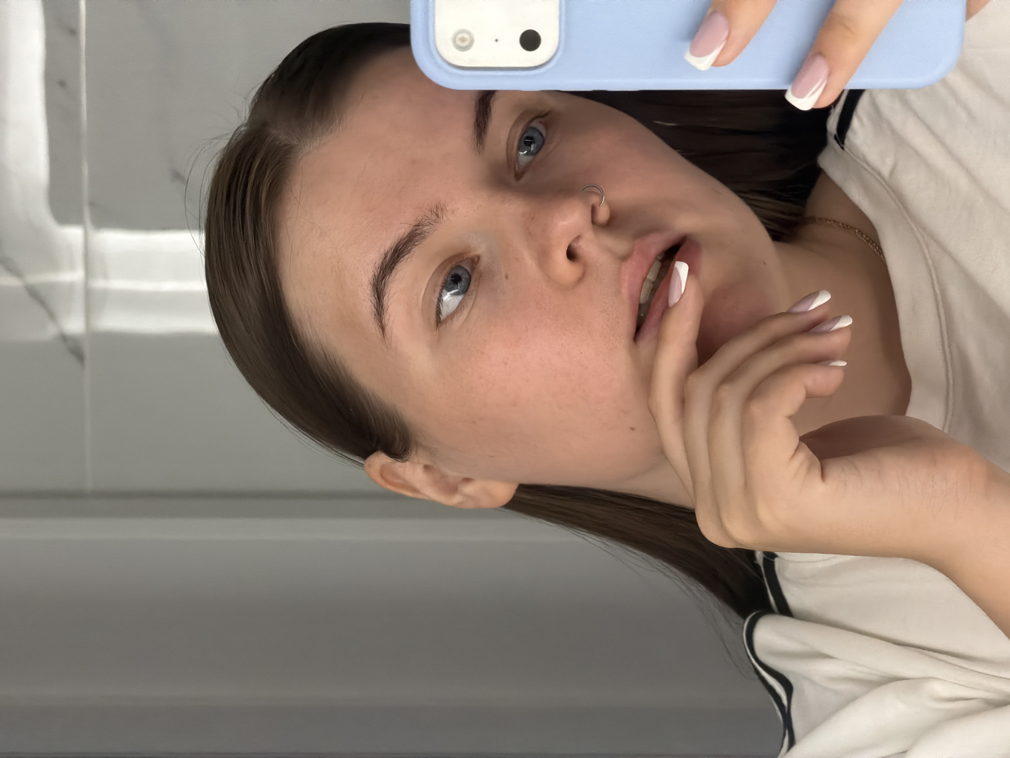 A young woman is taking a selfie in what appears to be a bathroom, as suggested by the plain white wall behind her. She is holding a light blue smartphone with a dual-lens camera in her right hand. She is gazing towards the camera with a slightly contemplative expression. Her hair is dark, and she has light-colored eyes and fair skin. A small, silver ring is visible in her left nostril. Her fingernails have a French manicure. She is wearing a white t-shirt with a black stripe along the shoulder. A delicate gold necklace is visible around her neck. Her fingers are lightly touching her lips. There is no contextual information to confirm the image was taken in Chaikovsky, Russia.