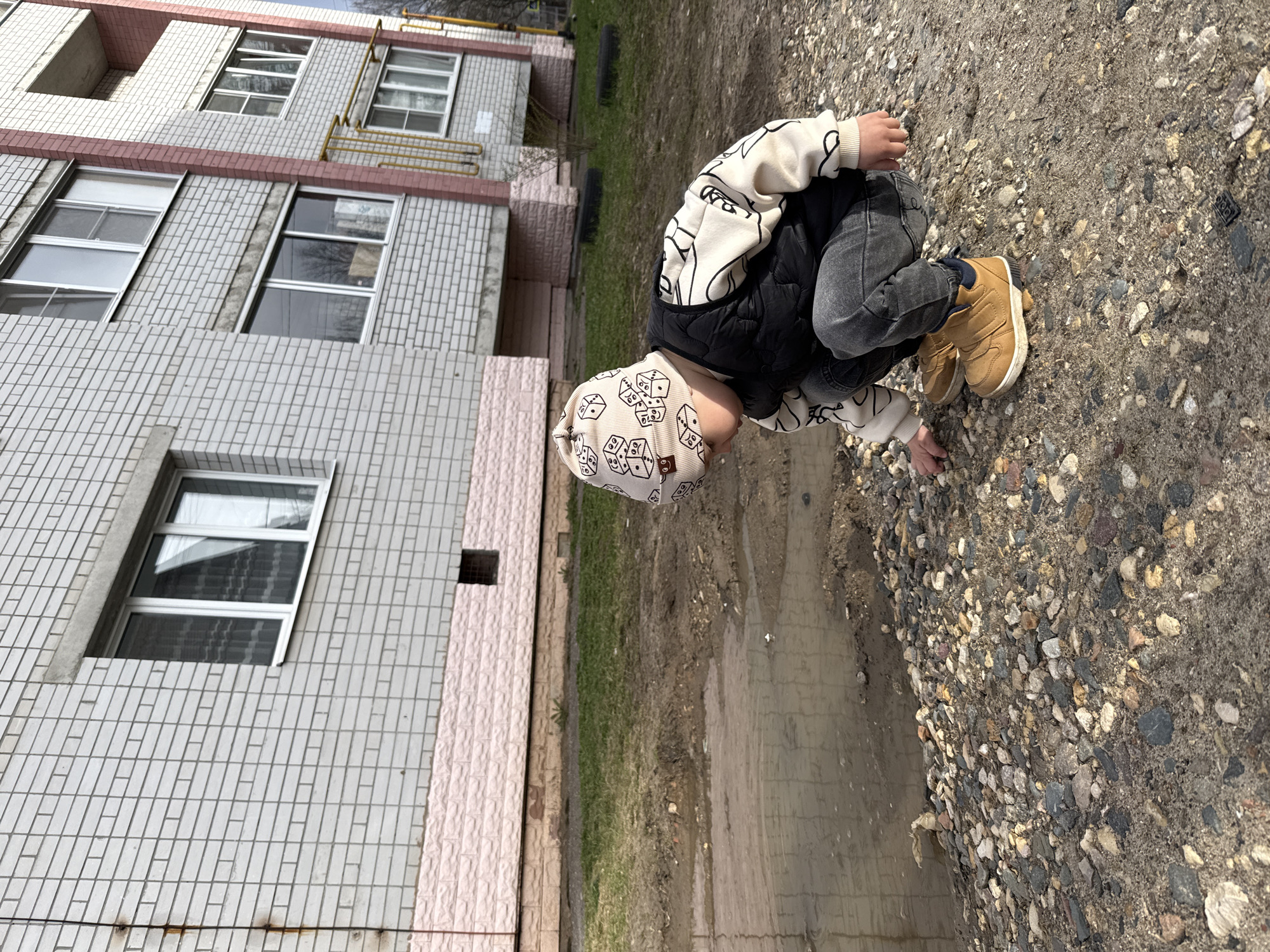 A young child wearing a beige hat with dice patterns, a black vest, and tan boots crouches near a puddle. The ground is a mix of gravel and dirt. Behind the child is a multi-story brick building with windows. The location is Rusanovo, Russia.