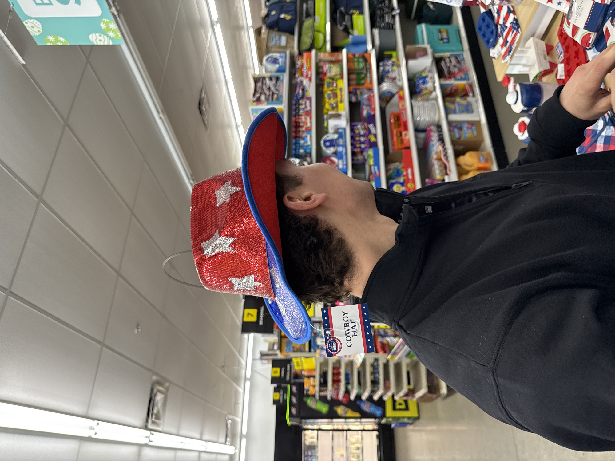 A man is wearing a red cowboy hat with white stars and a blue trim in a store. The hat has a tag attached that says "Cowboy Hat". The man is wearing a black jacket. Shelves filled with merchandise are in the background, indicating the location is a retail store. The photo was taken in Newport, United States of America.