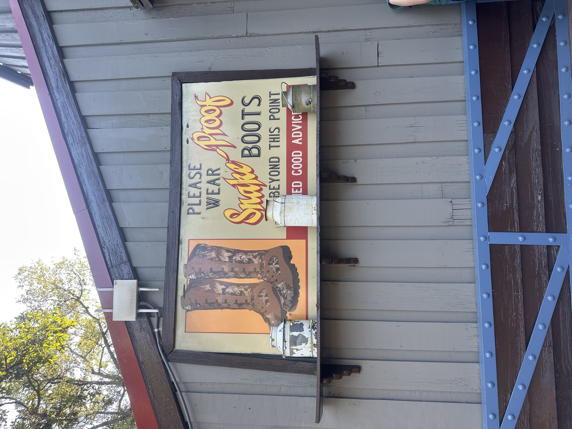 The image shows a vintage-style sign mounted on the exterior wall of a building. The sign is an advertisement for snake-proof boots, featuring the text "PLEASE WEAR Snake Proof BOOTS BEYOND THIS POINT ED GOOD ADVICE" along with an illustration of a pair of camouflage boots. The sign is framed and has metal props attached to it. The building's siding is painted a neutral color. To the right of the sign is a brown wooden structure with blue metal supports and rivets. The location is in Mexico City, Mexico.