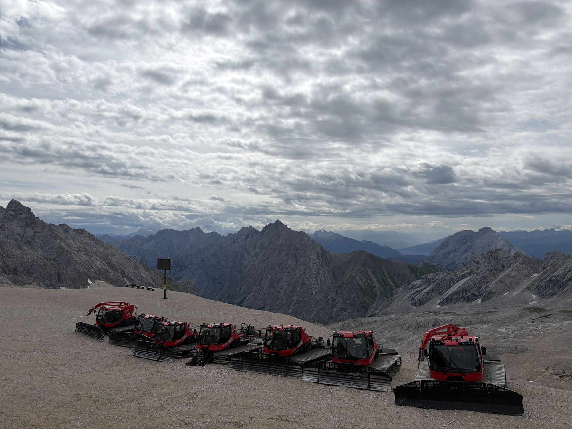 This is an eye-level, wide shot of a mountain landscape under a cloudy sky. In the foreground, there are seven red snow groomers parked in a line on a gravelly, rocky surface. The snow groomers have large caterpillar tracks and a wide blade in front. The mountains in the background are rugged and rocky, with some patches of snow. The sky is filled with a thick layer of gray and white clouds, with some lighter areas suggesting glimpses of blue sky. The overall scene conveys a sense of remoteness and high altitude. This scene is likely located on the Zugspitze, Germany's highest mountain, near Garmisch-Partenkirchen, Germany, as snow groomers are used to maintain ski slopes and trails in such mountainous regions.