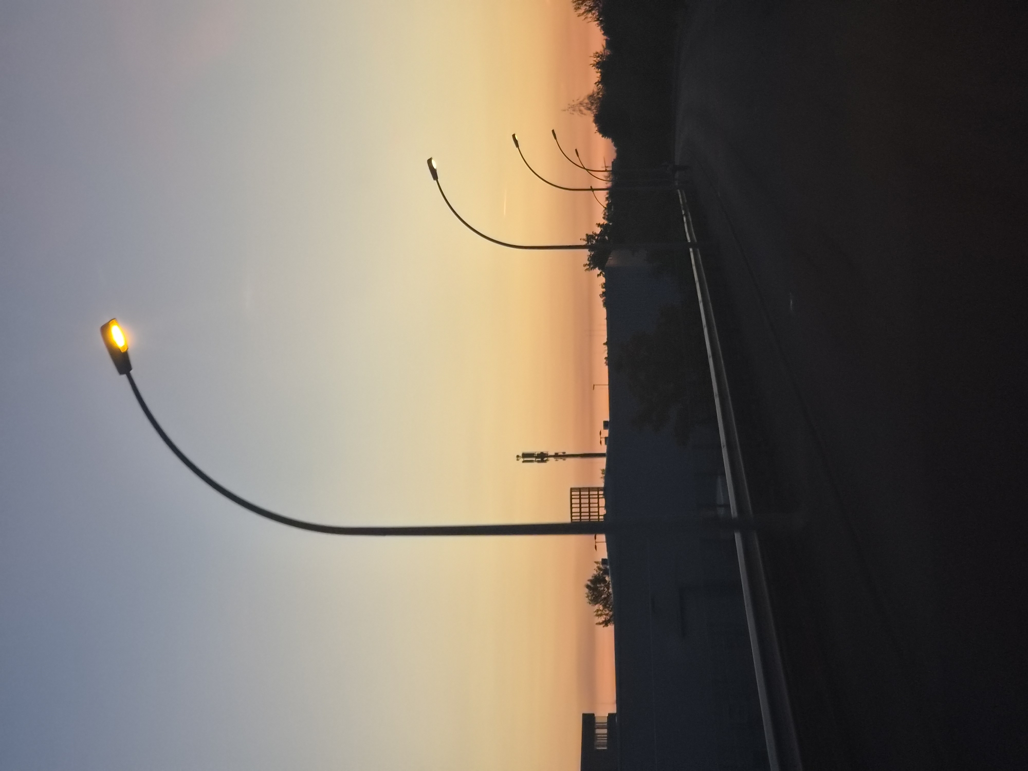 This image depicts a street scene at dusk in Leudelange, Luxembourg. Several modern streetlights with illuminated bulbs line the right side of a paved road, which curves into the distance. In the foreground, a dark shadow covers the road, with a reflective guardrail visible along the left edge. The background features the silhouette of a long, dark building, possibly an industrial structure or warehouse, and a telecommunications tower. A line of trees forms a dark horizon against the gradient sky, transitioning from pale blue at the top to warm orange and pink hues near the horizon. No people are clearly discernible in the image. The overall atmosphere is serene and twilight.