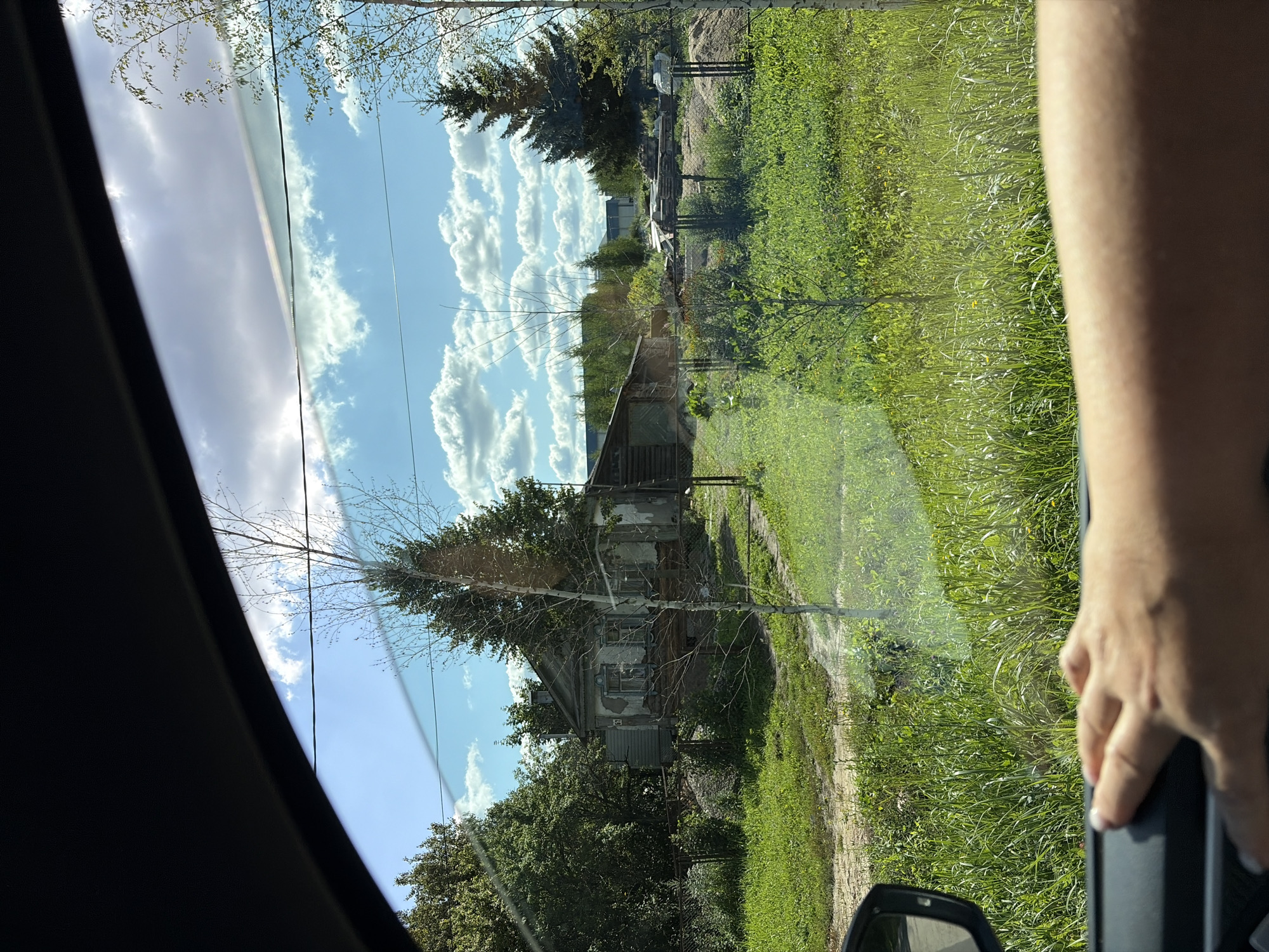 A hand rests on the dashboard of a vehicle, partially obscuring the view of a rural landscape through the car window. The scene outside depicts a weathered, old house with peeling paint and boarded-up windows, situated amidst overgrown green grass and trees. A small, sparsely leaved tree stands in the foreground. In the background, more trees and a larger evergreen tree are visible, along with what appears to be a long, low industrial building. The sky is a bright blue, dotted with fluffy white clouds, suggesting a sunny day. The location is identified as Novoye Podvyaznovo, Russia.