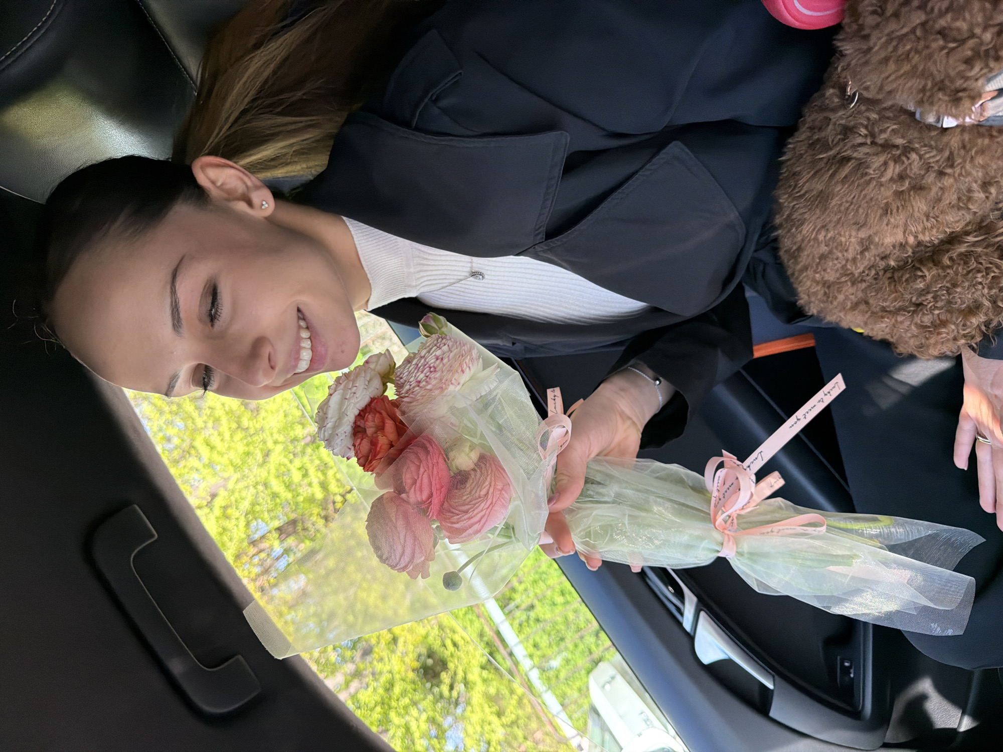 The image captures a joyful moment inside a car, featuring a young woman holding a bouquet of flowers. The woman, with her dark brown hair pulled back, is smiling broadly, looking towards the viewer with her head tilted. She wears a black jacket or trench coat over a white ribbed top, and a silver necklace is visible. Her visible ear features a small earring with three studs.
She is holding a delicate bouquet of flowers, appearing to be ranunculus, in shades of pink, white, and coral. The flowers are artfully wrapped in a light green translucent mesh and secured with a pink ribbon. The ribbon clearly displays the text "Lovely to meet you," repeated along its length.
To the right, partially visible behind her, is a fluffy, brown, curly-haired object, possibly a small dog or a plush toy. The interior of the car features black seating and a black overhead grab handle. Through the car window, bright daylight illuminates lush green foliage, indicating a sunny day during a season when trees are in full leaf, likely spring or summer. The scene conveys a sense of happiness and appreciation. The location context for this image is Sofia, Bulgaria.