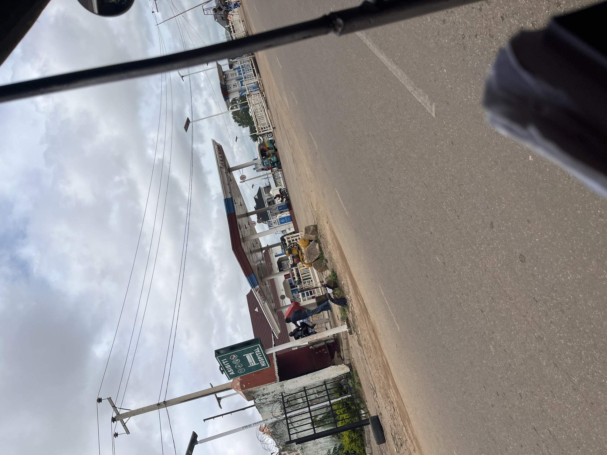 This is a photograph taken from a vehicle in motion, likely a tricycle, in Rafin Par, Nigeria. The scene depicts a gas station with multiple pumps under a covered canopy. In the foreground, to the left, a green sign with white lettering reads "ASIBITI HOSPITAL" accompanied by four red icons that resemble a heart with a cross, suggesting medical services are available nearby. A black metal fence encloses the area behind the sign. Two individuals are visible near the entrance of a building adjacent to the gas station. One is seated on a low wall, while the other stands and appears to be interacting with a red object. Further into the gas station, a tricycle is parked next to a pump. The road in front of the gas station is paved, with a white line marking the lanes. The sky is overcast with clouds, and electrical poles with wires are visible in the background. The overall impression is of a typical roadside scene in a Nigerian town.