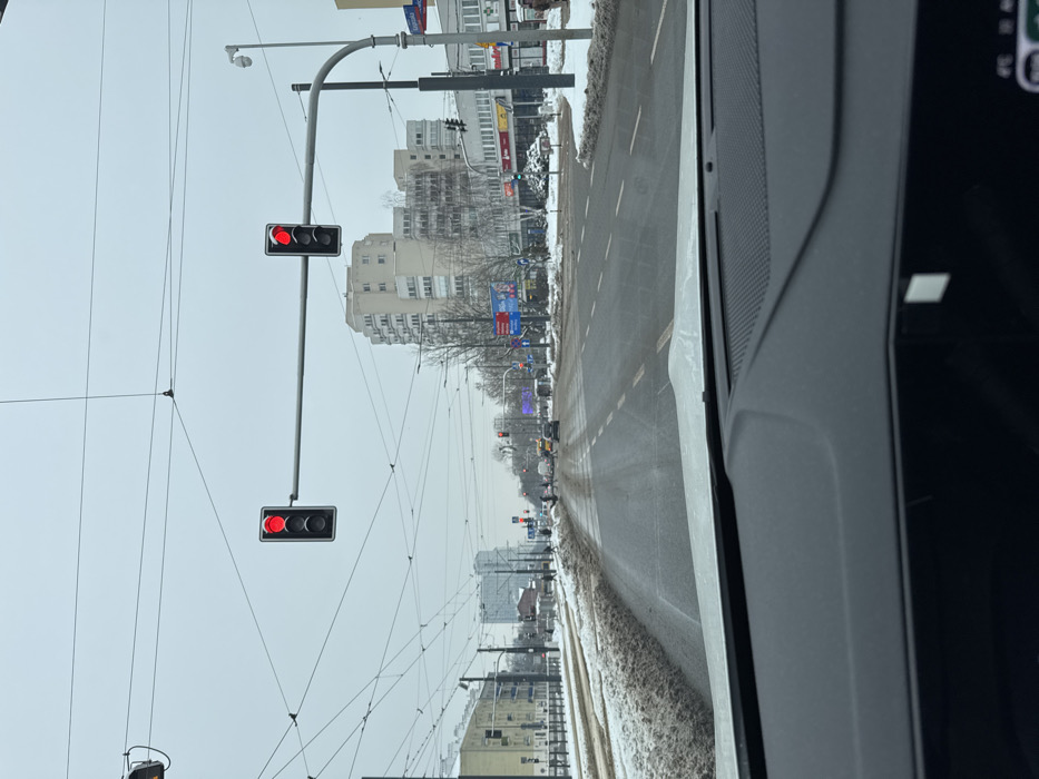 The image captures a winter street scene from inside a vehicle at the intersection of Grójecka Street and Kopińska Street in Warsaw, Poland. The lower foreground shows the dark dashboard of the vehicle.
The sky is uniformly overcast and light gray. The multi-lane asphalt road extending into the distance is wet and appears slushy, with visible tire tracks. Accumulated dirty, brownish snow and slush form piles along the road edges and a median strip to the left, which also contains exposed tram tracks. Multiple overhead wires, likely for trams and utilities, crisscross the sky. Two primary traffic lights suspended horizontally across the road display active red signals, indicating a stop. A blue street sign on a pole to the right identifies "ul. Grójecka" and "ul. Kopińska."
In the mid-distance, several pedestrians are observed utilizing a crosswalk. Multiple vehicles, including a light-colored taxi, are stopped at the intersection. On the right side of the street, a prominent multi-story, light-colored building with balconies is visible behind a row of street-level commercial establishments. Buildings and partial scaffolding are also present on the left side of the street further down the road. A street lamp and a security camera are mounted on the pole on the right.