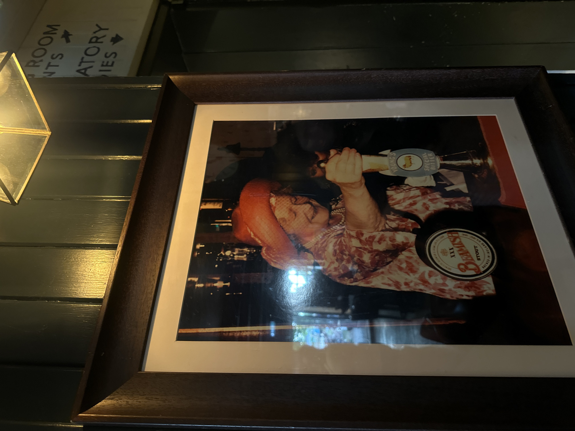 The image shows a framed photograph hanging on a dark green, wooden paneled wall. To the left of the frame, a geometric light fixture with a golden frame is visible. Above the frame, a white sign with black lettering is partially visible, displaying the words "ROOM" and "ATORY" with arrows pointing in different directions.
The framed photograph depicts an individual (possibly a woman) in a red hat and floral patterned clothing, interacting with a beer tap. The tap handle is labeled "YOUNG'S SPECIAL BITTER". A "Beamish Stout" tap marker is also visible. The photograph appears to be taken in a pub or bar setting.