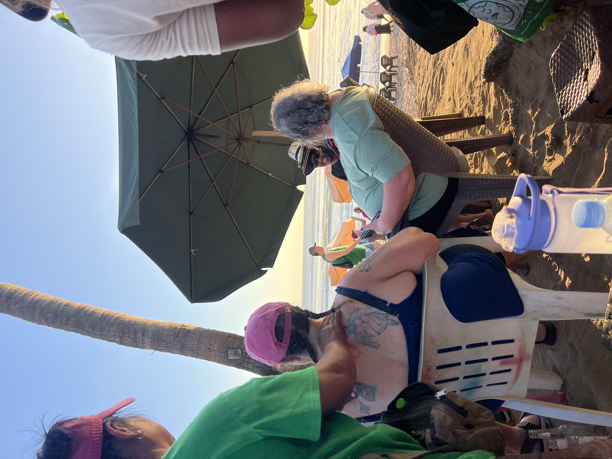 The image depicts a beach scene in Kuta, Indonesia, during sunset. In the foreground, a person in a green t-shirt is giving a back massage to another person seated in a white plastic chair. The person receiving the massage has a pink baseball cap and a blue bathing suit, and their back is adorned with several tattoos, including a prominent turtle. The beach is sandy, and a large green umbrella provides shade. In the mid-ground, other people are visible relaxing on chairs and walking along the shore, with the ocean and a clear sky in the background. The lighting suggests it is late afternoon or early evening.