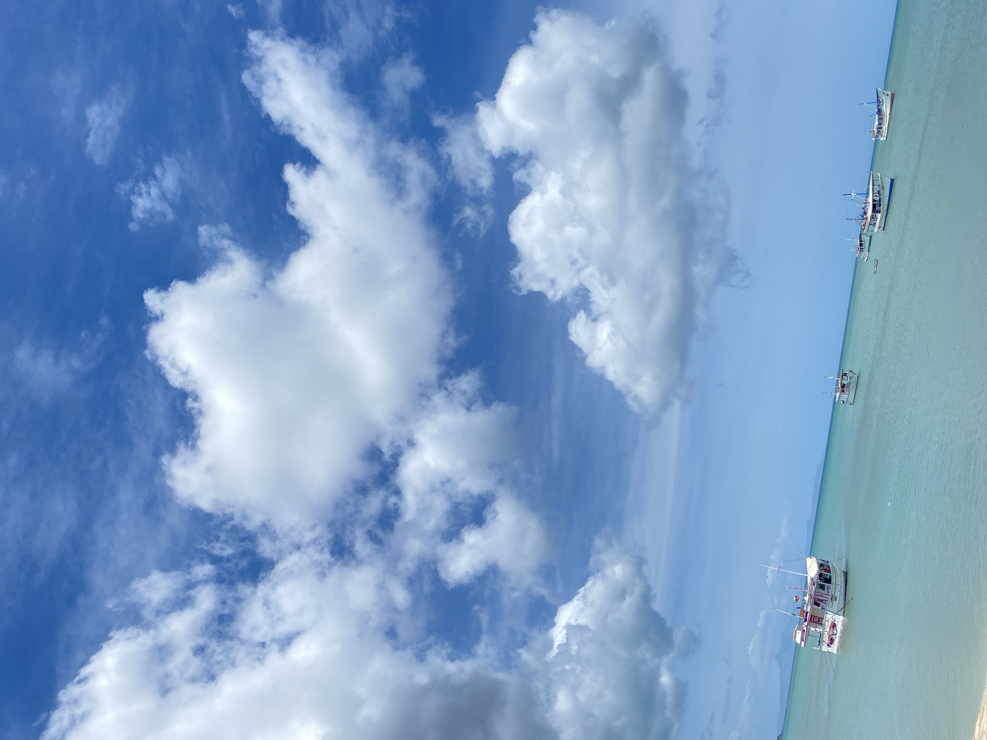 The image depicts a scene in Juan Griego, Venezuela, showing a bright blue sky with scattered white clouds. A body of light blue water is visible, with several white boats floating on the surface. There are no people visible in the image.