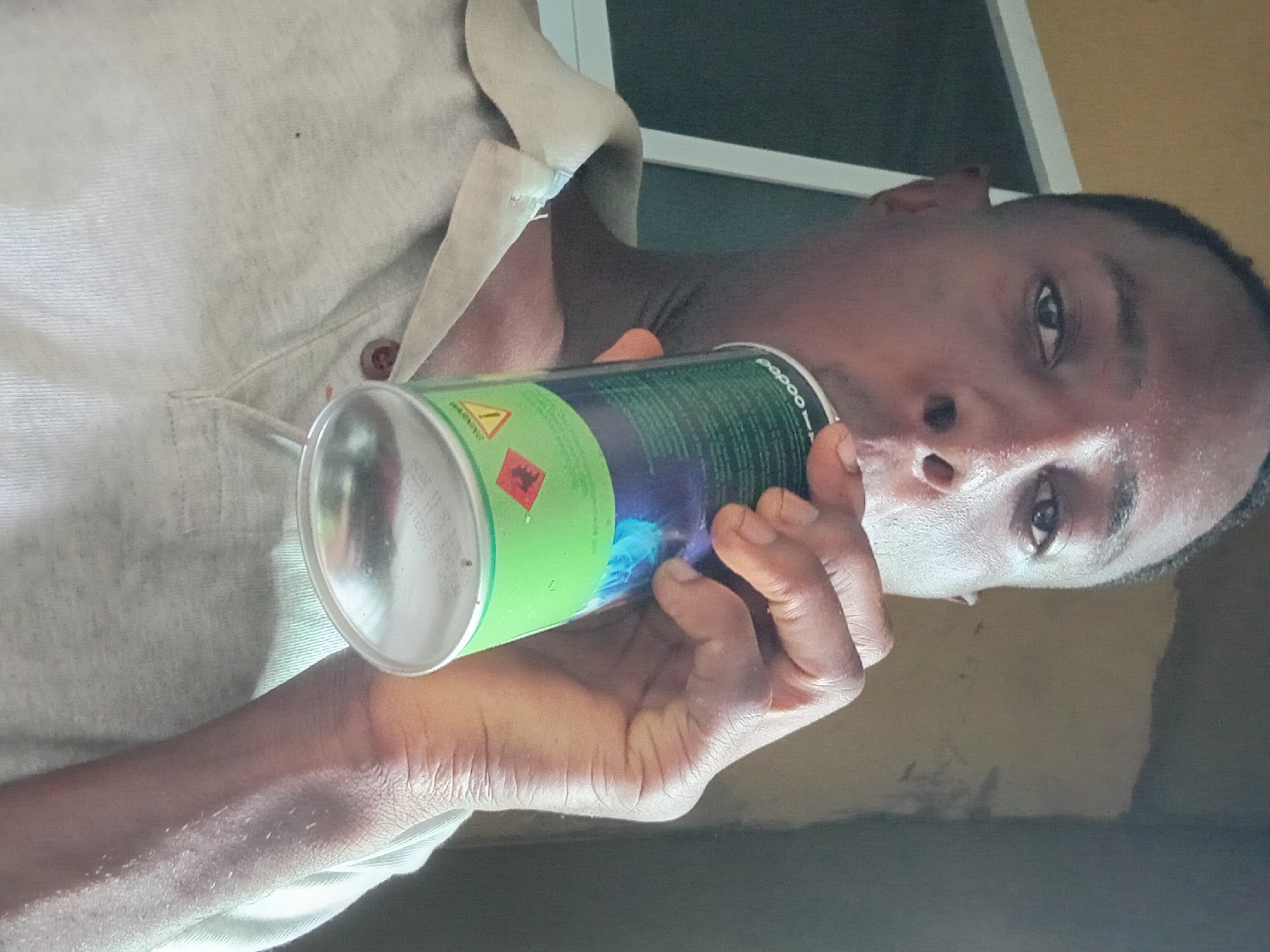 An adult male is holding a green aerosol can up to his face. The can has a red flame symbol on it. The man is wearing a light-colored collared shirt. The location is Akwete, Nigeria.