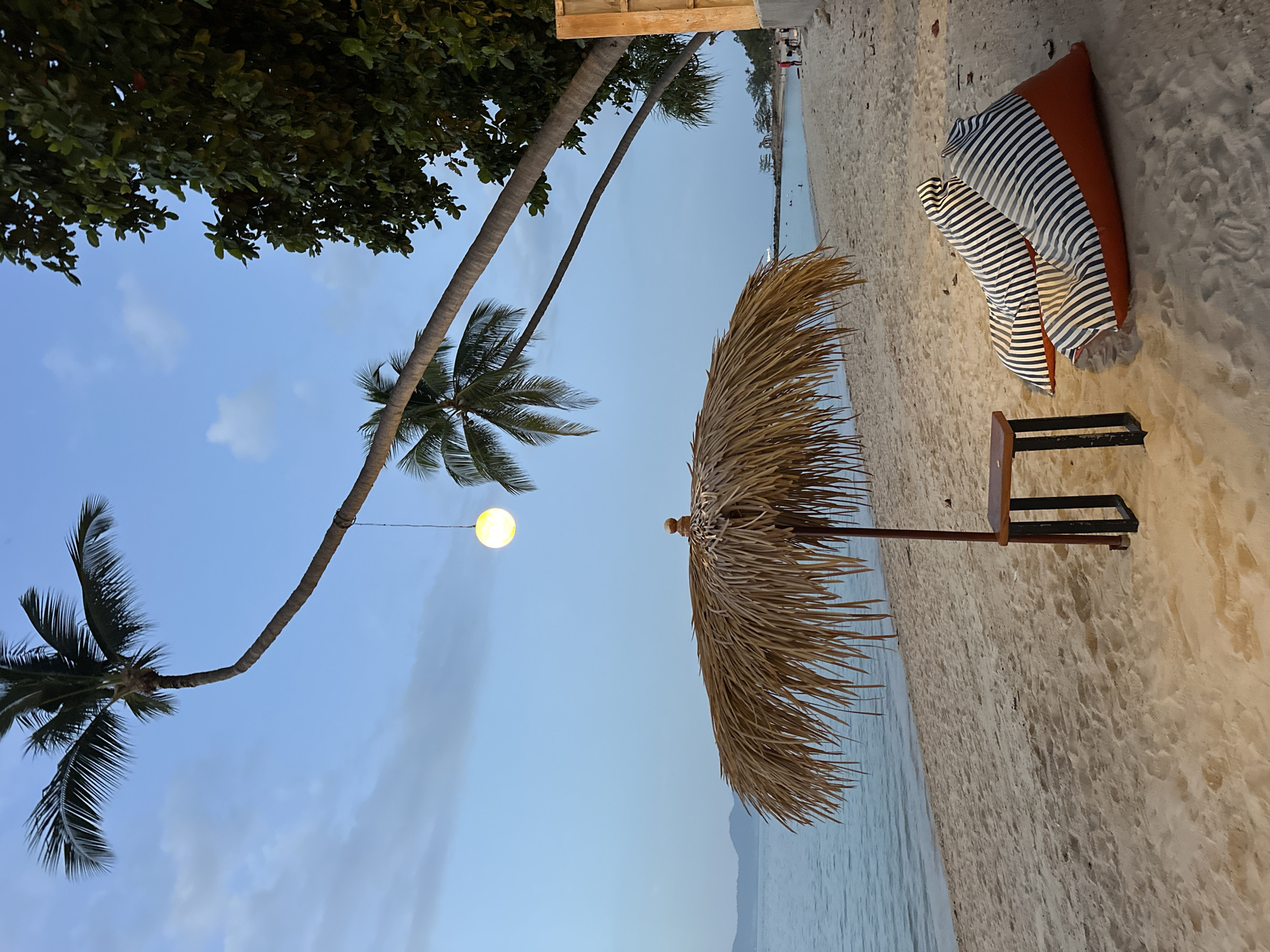 On a sandy beach in Ko Samui District, Thailand, the scene unfolds at dusk under a pale blue sky. Two large, striped beanbag chairs, one orange and one with orange and white stripes, rest on the sand near a small wooden table with black metal legs. A thatched umbrella provides shade, its dry fronds casting a textured shadow. Two palm trees lean gracefully over the beach, their fronds silhouetted against the sky, and a single, round, illuminated lantern hangs suspended between them. The calm ocean stretches to the horizon, with a faint outline of distant hills. The sand shows footprints, suggesting recent activity. In the background, a few people are visible near the water's edge and further down the beach, engaged in various leisurely activities. The overall atmosphere is serene and relaxing, with the natural beauty of the beach and tropical vegetation illuminated by the soft evening light and artificial lantern.