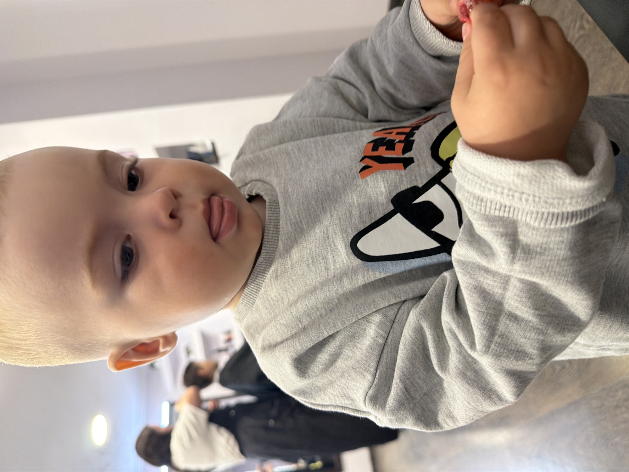 A young child is prominently featured in the image, sticking their tongue out slightly. They are wearing a gray long-sleeved shirt with a cartoon design. In the blurred background, two adults are visible. Based on the artifacts of the image, it is not possible to confirm that this photo was taken in the city of Votkinsk, Russia.