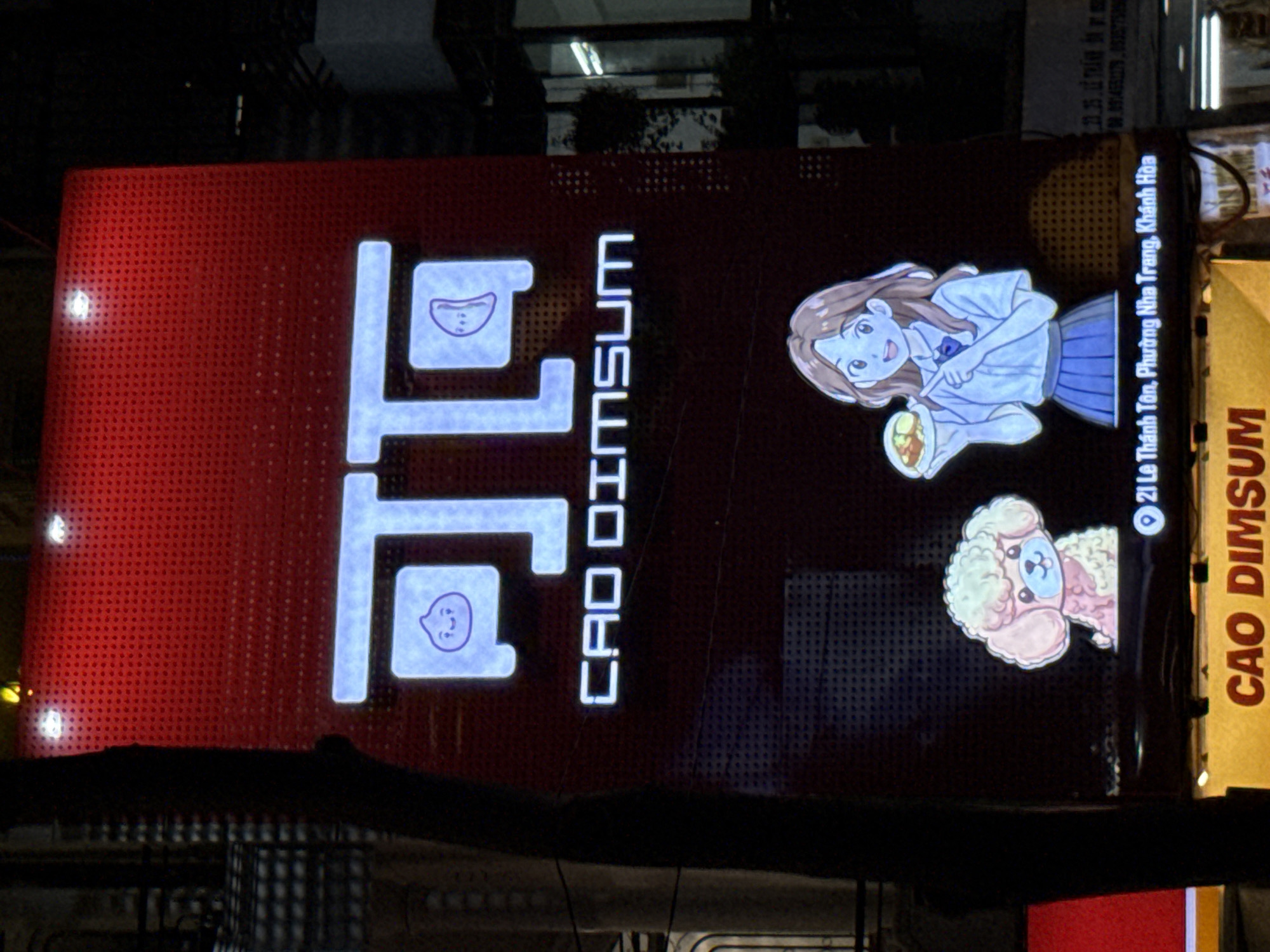 The image shows a brightly lit red sign for "CAO DIMSUM" at night, located at 21 Le Thanh Ton, Phuong Nha Trang, Khanh Hoa. The sign features a stylized logo with two blue characters resembling dumplings with faces, and the text "CAO DIMSUM" in white lettering. Below the logo, there are cartoon illustrations of a pink poodle and a young woman in a school uniform holding a plate of food. The background is dark, suggesting an outdoor setting at night. The sign appears to be a storefront or a restaurant advertisement.