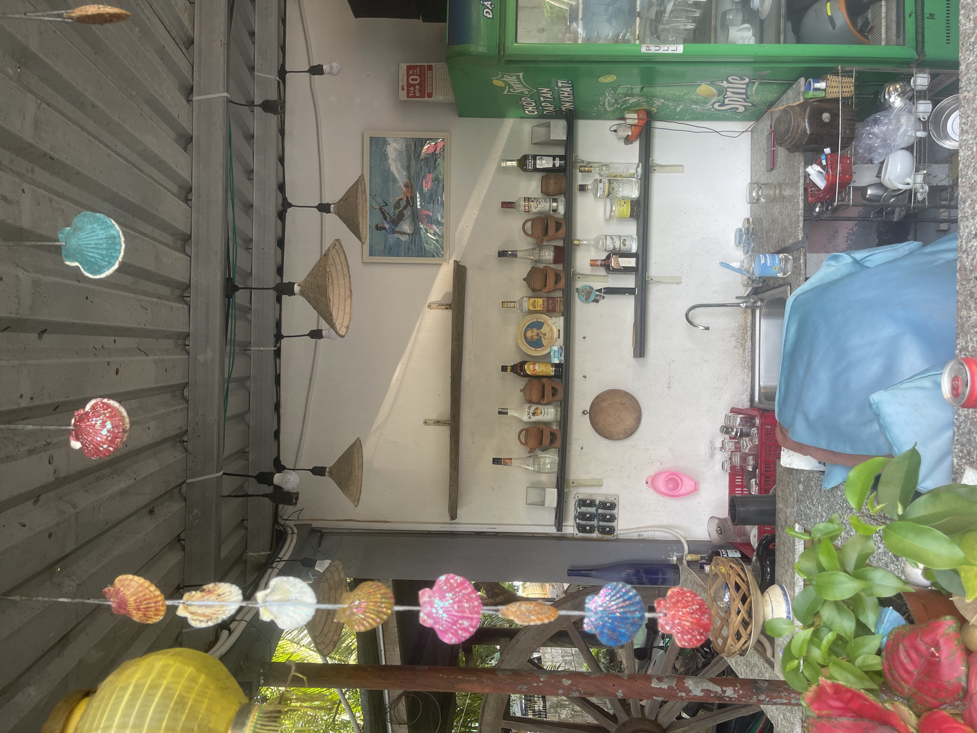 The image shows an outdoor bar or food stall in Phan Thiet Ward, Vietnam. The bar is situated on a concrete counter with a stainless steel sink and faucet on the left. Behind the counter are shelves displaying various bottles of alcohol, some ceramic pots, and decorative items. A green Sprite refrigerator is on the right side of the frame. Hanging from the ceiling are strings of colorful seashell decorations and straw lamps. A large framed picture of a person kitesurfing hangs on the wall above the shelves. In the foreground, there are plants and a blue tarp covering a portion of the counter. A red and white soda can is visible on the lower right. The overall impression is of a casual, open-air establishment, likely catering to tourists or locals seeking refreshments in a tropical setting.