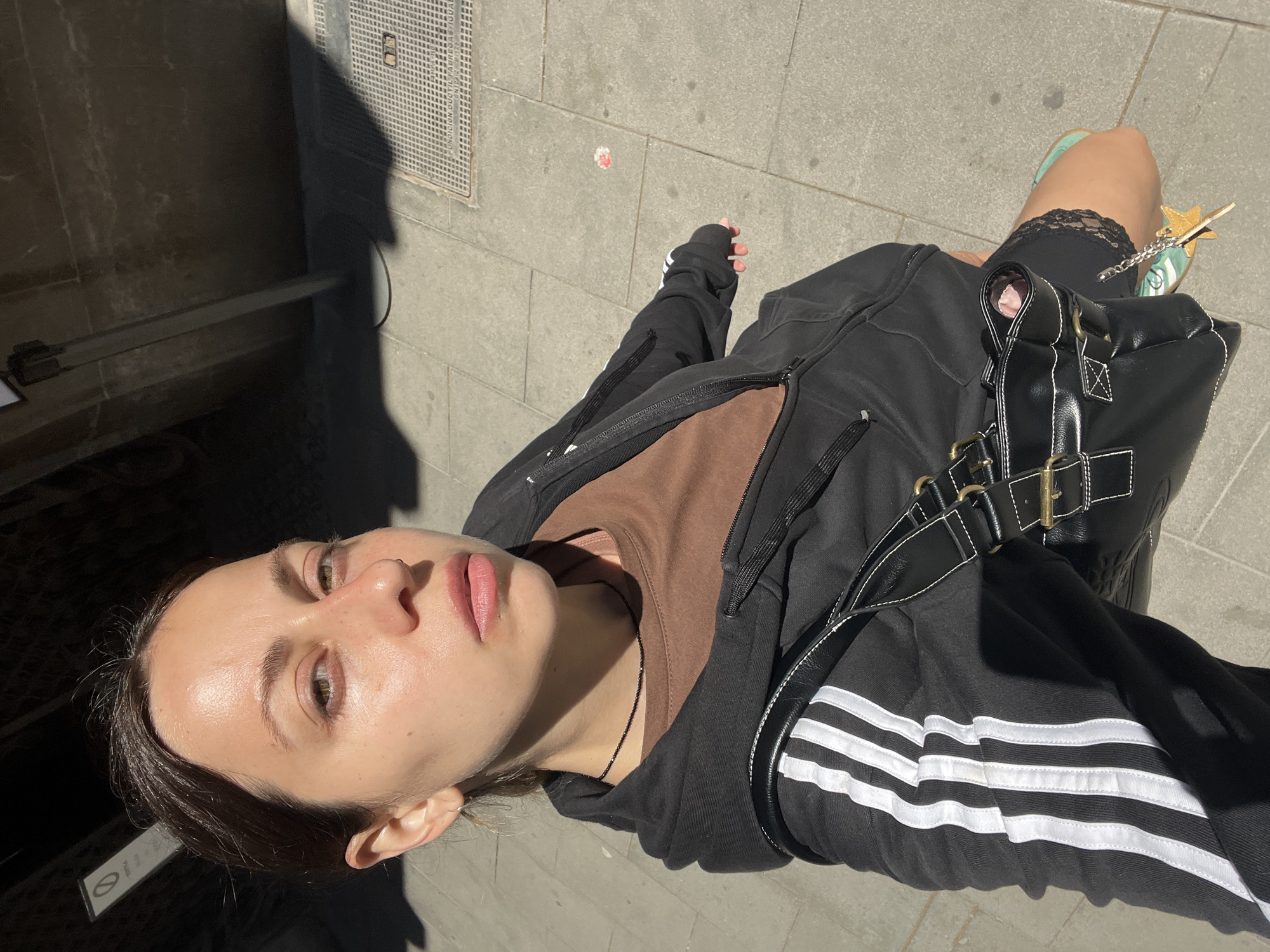 A high-angle, top-down outdoor selfie captures a young woman looking directly upwards at the camera on a bright, sunny day. The strong sunlight creates distinct shadows on the grey rectangular paving stones, including a large silhouette of a person to the right, suggesting the photographer.
The subject has dark hair, light skin, and green/hazel eyes, with a neutral or slightly contemplative expression. She wears a black zip-up hoodie with white Adidas stripes on the left sleeve, layered over a brown t-shirt, and a thin black necklace. Black shorts with a delicate lace trim are visible on her right thigh. She carries a black shoulder bag with white contrast stitching and gold buckles. Her right foot, partly visible, wears a turquoise shoe adorned with a yellow star-shaped charm.
In the background, a metal grate on the pavement displays partially legible text: "FUNDICION DUCTIL FABRICADA GU...". To the far left, a "no entry" sign (a white circle with a diagonal line) is visible on a darker surface. The setting appears to be an urban environment, consistent with the provided context of Barcelona, Spain.