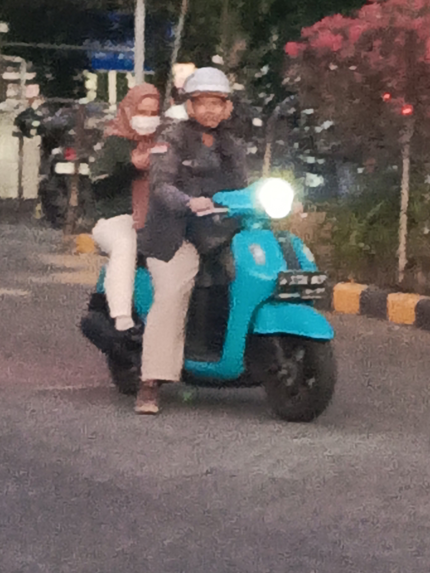 This is a slightly blurry, medium-shot, eye-level, outdoor, nighttime photograph of two individuals riding a bright turquoise scooter. The scooter has its headlight on, illuminating the foreground. The driver, a male wearing a white helmet, appears to be looking forward. He is wearing a dark jacket and light-colored pants. The passenger, a female, is seated behind him and is wearing a light brown hijab and a white face mask. She is wearing white pants. The scooter is on a paved road, likely in Sidoarjo, Indonesia, as indicated by context. In the background, there are trees with red and pink blossoms, a street light pole, and what appear to be concrete barriers with yellow and black markings. Other vehicles are partially visible in the background, suggesting this is a street or a parking area. The overall scene is dimly lit.