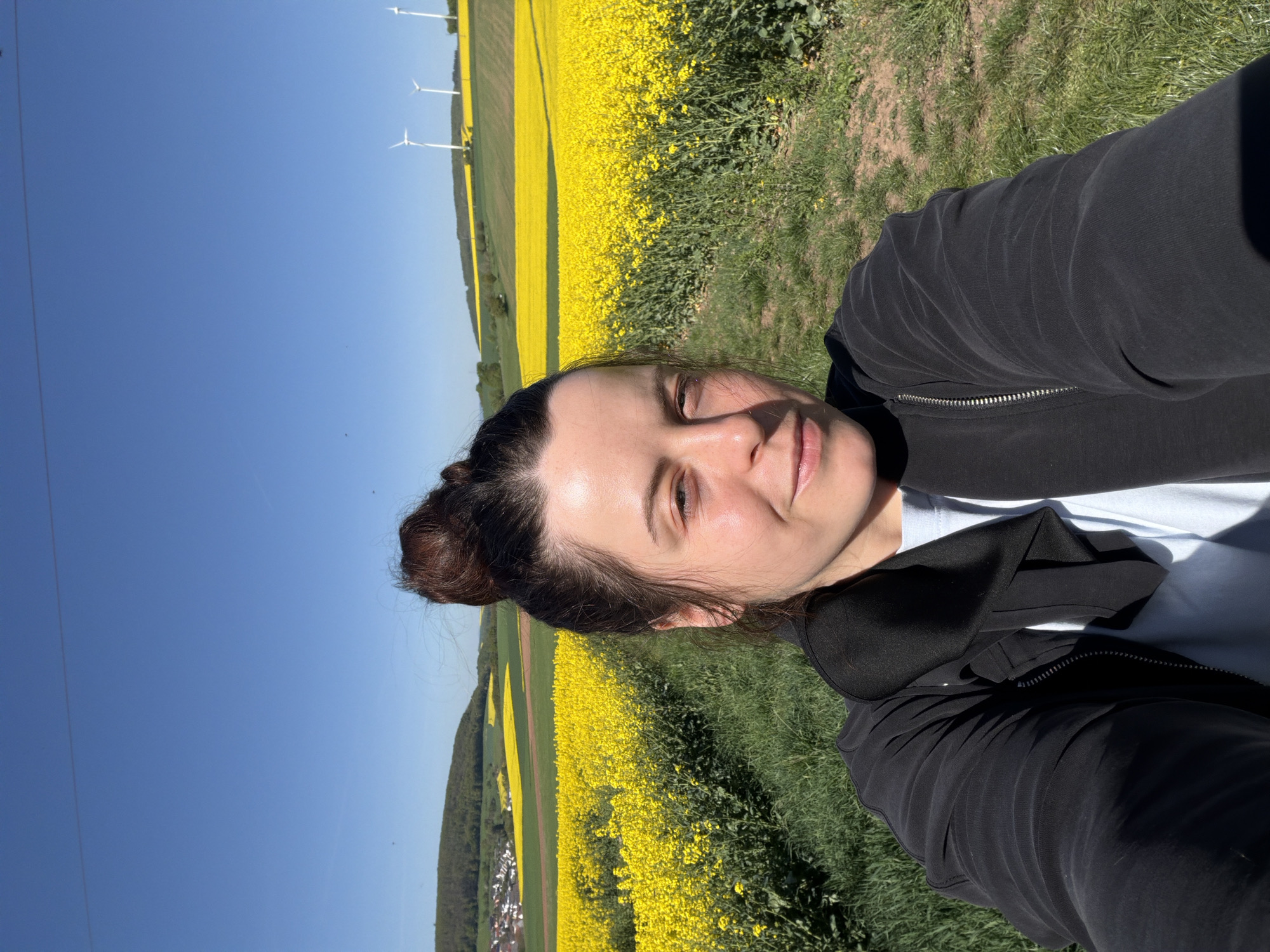 A selfie-style portrait captures a woman outdoors against a vibrant rural landscape near Obermoschel, Germany, on a clear, sunny day. The woman, with dark hair tied back in a bun and a slight smile, looks directly at the camera. She is wearing a black zipped hoodie over a light-colored top.
Behind her, the scene is dominated by vast fields of bright yellow flowers, indicative of blooming rapeseed or canola, extending across gentle hills. These yellow fields are interspersed with strips of green vegetation and darker soil. In the distance, several tall white wind turbines stand against the expansive blue sky, and a small town or village is visible nestled on a distant hillside. The bright sunlight suggests it is daytime, and the clear sky indicates pleasant weather. No visible text is present in the image.