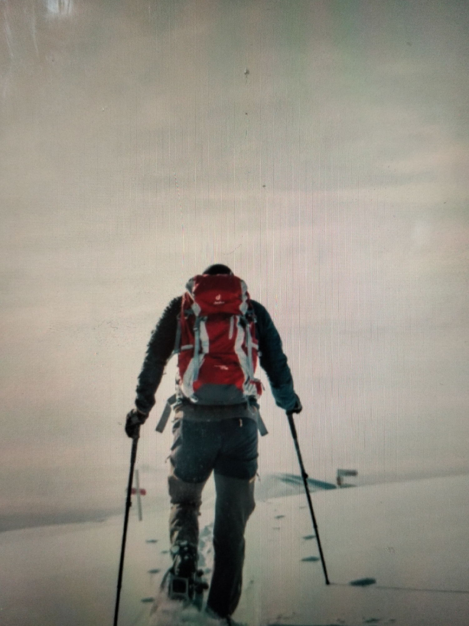 A person is backcountry skiing in Payagaboh, Indonesia. The person is wearing a red backpack, dark jacket, and grey pants. They are holding ski poles and appear to be moving forward. The environment is snowy and appears to be a winter landscape.
