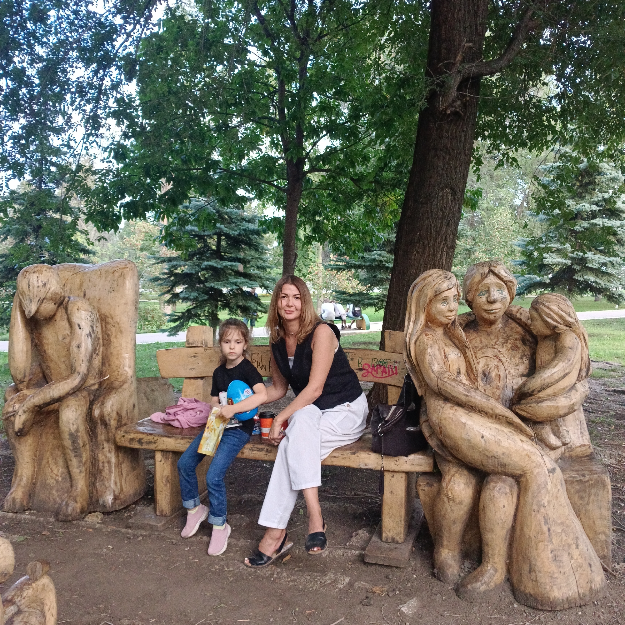In Samara, Russia, a mother and daughter are seated on a wooden bench in a park. The mother, a woman with blonde hair, wears a black vest over a white top and white pants. She is smiling and looking at the camera. Her daughter, a young girl with brown hair, is wearing a black t-shirt and blue jeans. She is holding a blue ball in her left hand and a yellow object in her right hand. She is looking towards the left side of the frame. The bench is flanked by two wooden sculptures. On the left is a sculpture of a seated figure with its head in its hands. On the right is a sculpture depicting three figures, two embracing a central figure. The park setting features trees and greenery in the background.
