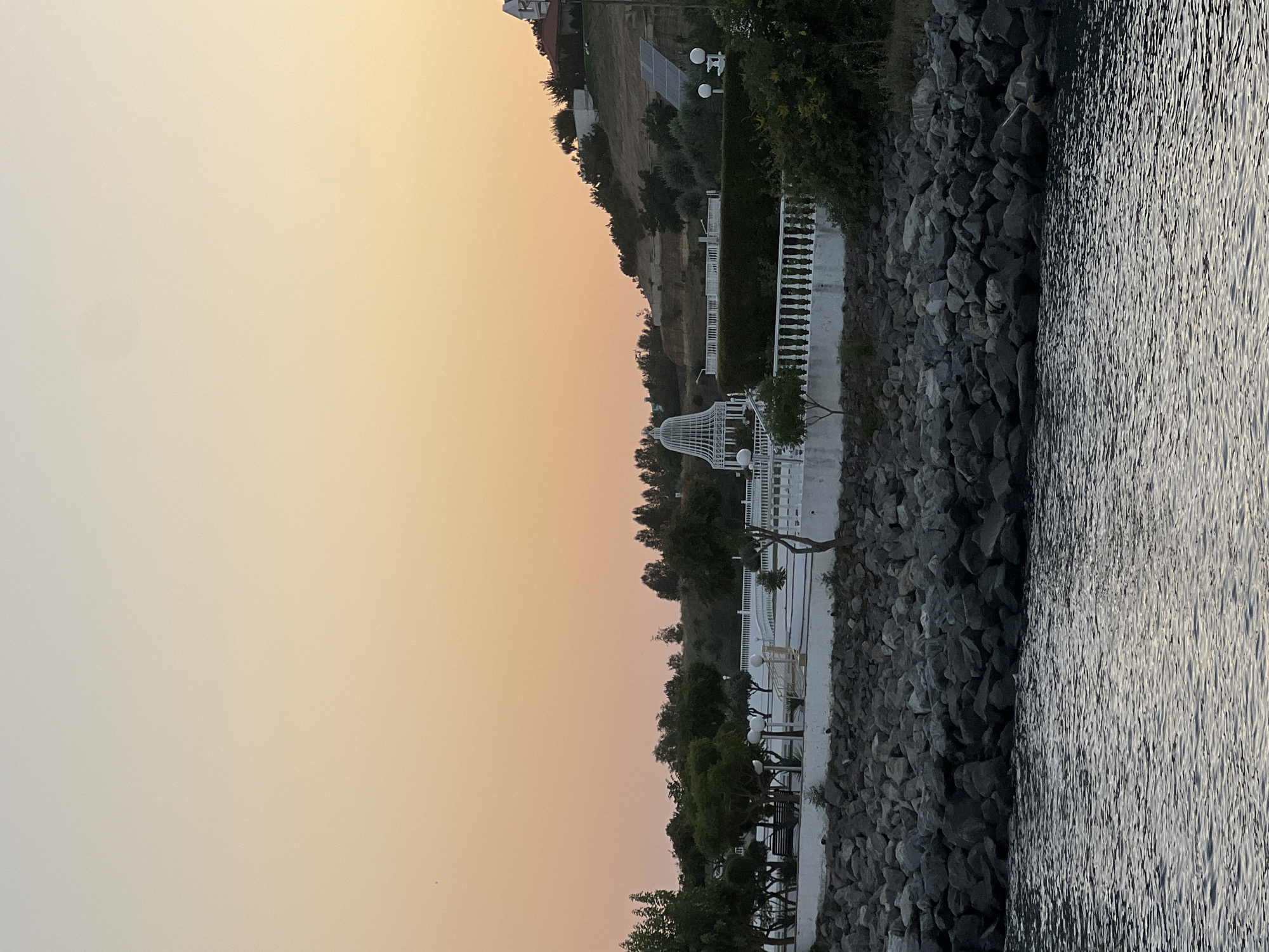 The image depicts a waterfront scene at dusk, possibly in an urban or resort area. In the foreground, a body of water with ripples is visible, suggesting it is a calm sea or large lake. Beyond the water, a rocky shoreline lines the edge of the land.
The land area features a white ornamental fence and several trees and bushes. Behind the fence, a white gazebo-like structure stands out, as does a paved walkway or patio area. Further inland, the terrain slopes upwards, with more trees and vegetation covering the hillside. A building with a red roof is visible on the upper part of the hill, and solar panels are also present on the slope.
The sky is a gradient of soft orange and yellow hues, characteristic of sunset or sunrise. No people are visible in the image. The overall impression is one of a tranquil, possibly developed, waterfront landscape.
