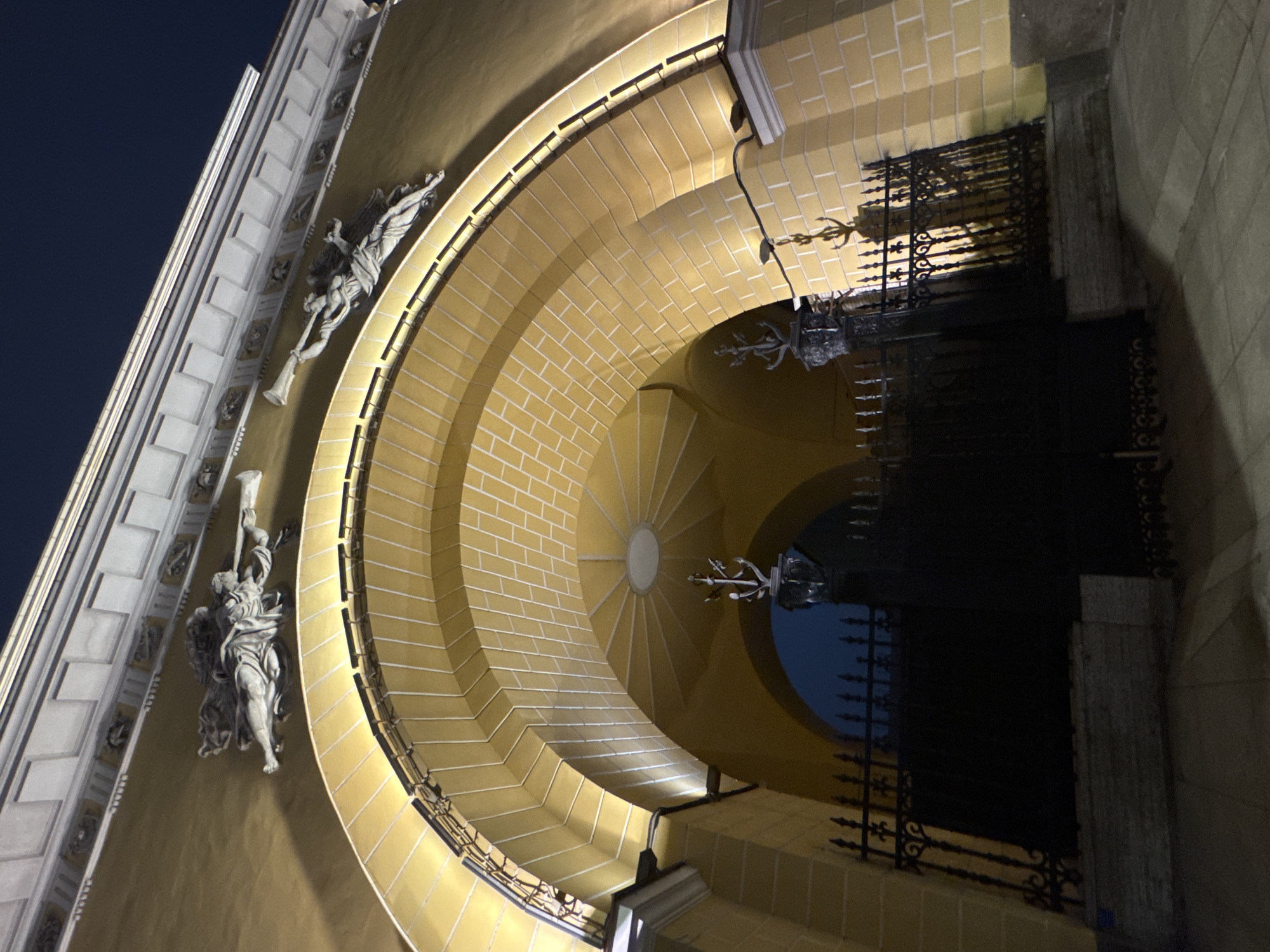 The image shows an exterior view of a building at night in Saint Petersburg, Russia. The building's architectural details include a semi-circular archway constructed of light yellow bricks, adorned with ornate light fixtures, and a black wrought iron gate. Sculptural figures, possibly angels, are mounted on the building's facade. The sky is visible through the archway, transitioning from dark blue to black. The image is angled upwards, giving a dynamic perspective of the structure.
