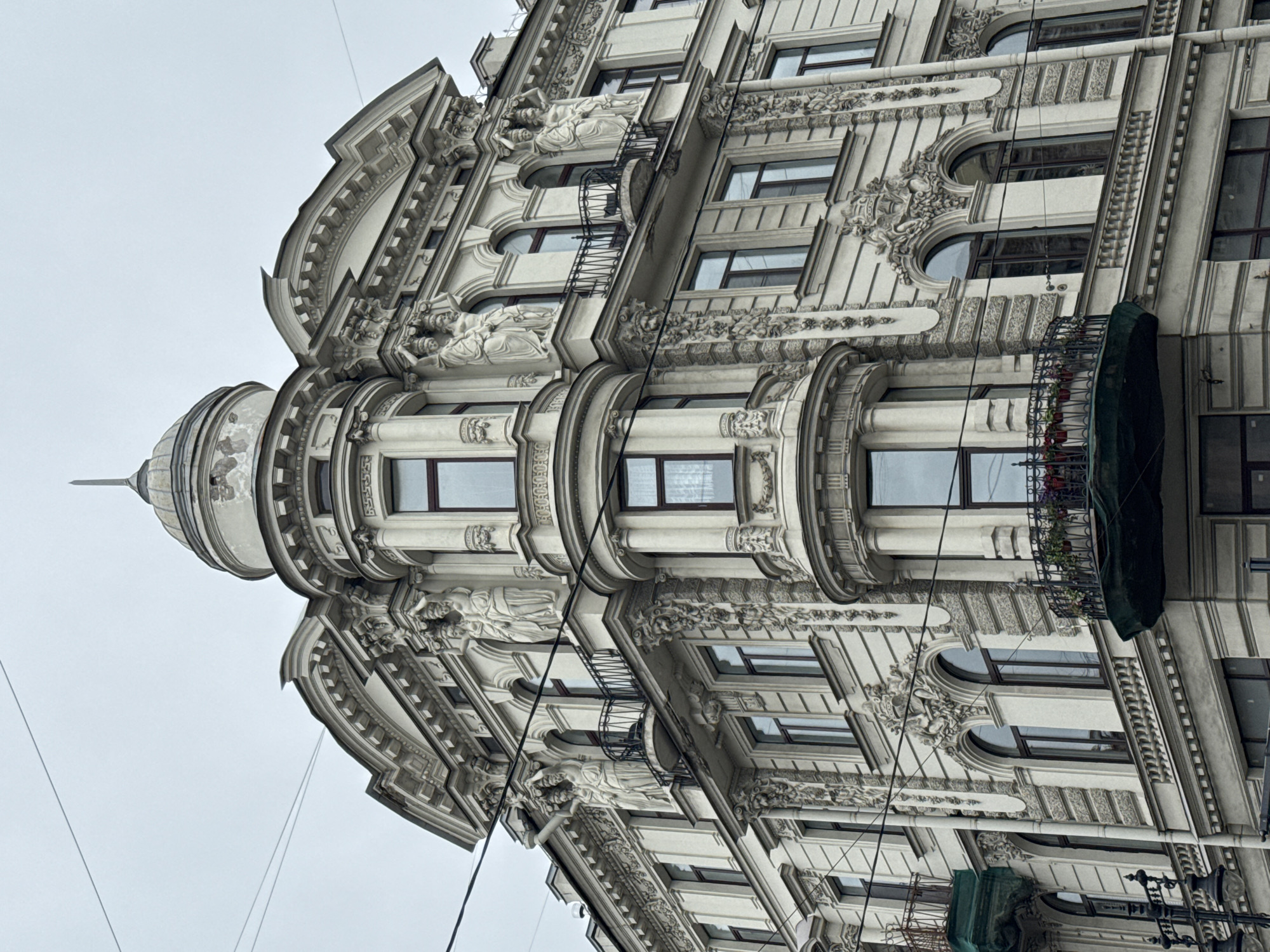 Here is a description of the image:
The image shows a corner building in Saint Petersburg, Russia, displaying elaborate architectural ornamentation. The building's facade is predominantly white or light gray, featuring rows of windows with ornate frames and decorative elements. The corner section of the building is rounded, topped with a dome-like structure and a pointed spire. The architectural details include sculpted figures, floral patterns, and geometric designs. Balconies with wrought-iron railings are present on some floors, and power lines run across the front of the building. The sky is overcast, creating a muted light across the scene.