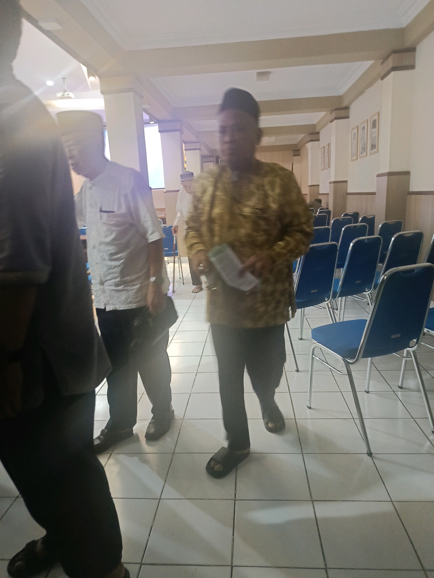 A group of men are walking and standing in a room with tiled floors and rows of blue chairs. The man in the foreground is wearing a gold patterned shirt and dark pants. He is holding a white booklet in his left hand. To his left, another man in a white shirt and dark pants is also walking, carrying a dark bag. In the background, more men are visible, some standing and some seated. The room appears to be a meeting or event space, with framed pictures on the wall. The lighting suggests it is indoors, possibly during the day. The scene appears to be located in Yogyakarta, Indonesia, given the context provided.