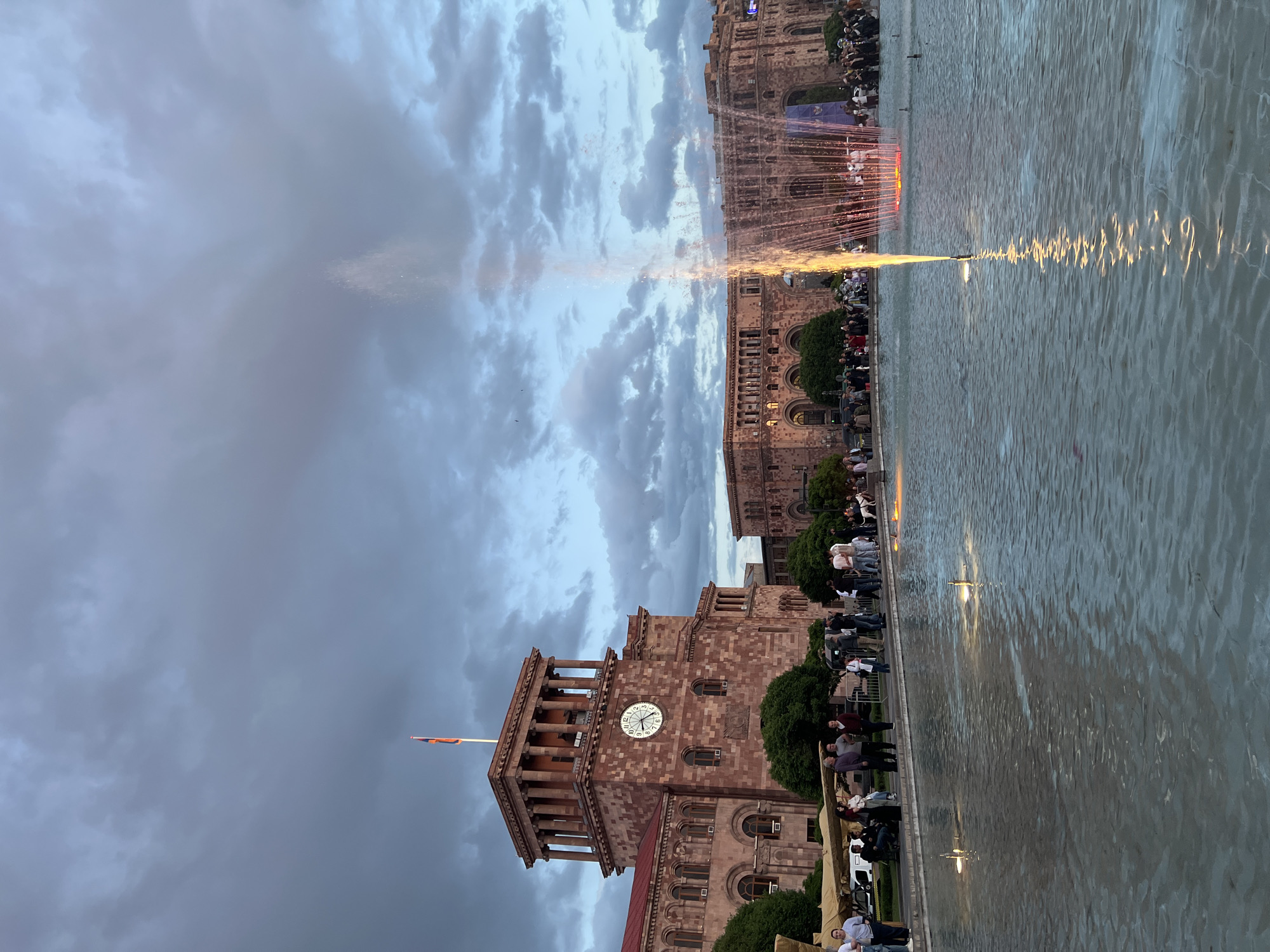 The image captures a scene in Yerevan, Armenia, focusing on Republic Square. The foreground features a large pool of water, likely part of the square's fountains, with water shooting upward. Along the pool's edge are many people, possibly tourists and locals, gathered to watch the fountain display.
In the background, the building is constructed from a pink-orange stone. There is a clock tower attached to the building. The sky is filled with heavy, dark clouds, giving the scene an overcast and dramatic feel. The image is vertical.