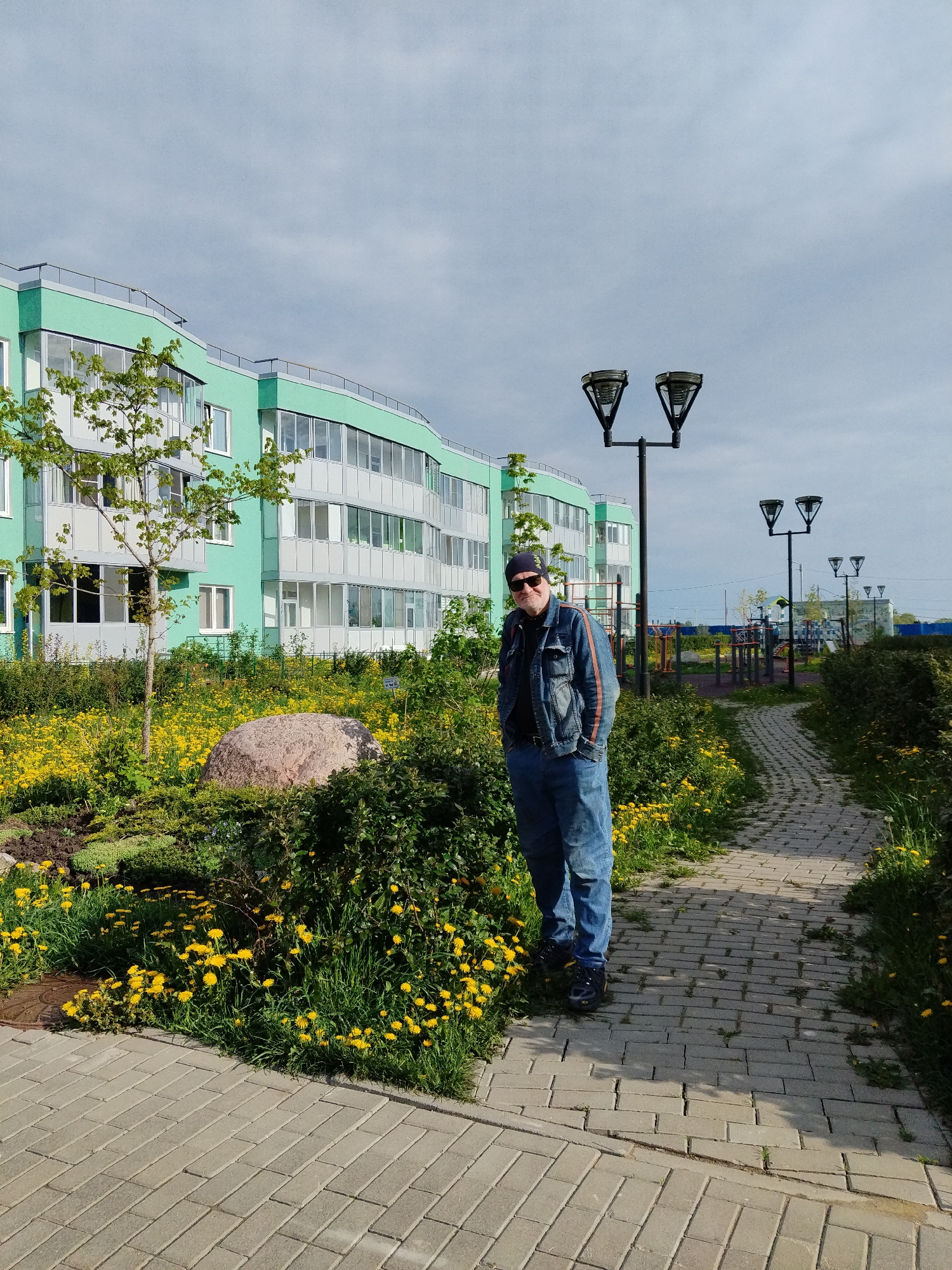 The image shows a man standing on a brick pathway in Kommunar, Russia. He is wearing a denim jacket and jeans, with sunglasses and a dark beanie. He has his hands in his pockets. To his left is a landscaped area with a large rock, bushes, and yellow flowers. Behind this area is a green and white building. To the right of the man, the brick pathway extends into the distance, lined with bushes and flowers, and lit by street lamps. In the background, beyond the pathway, is a playground. The sky is overcast with a mix of blue and gray.